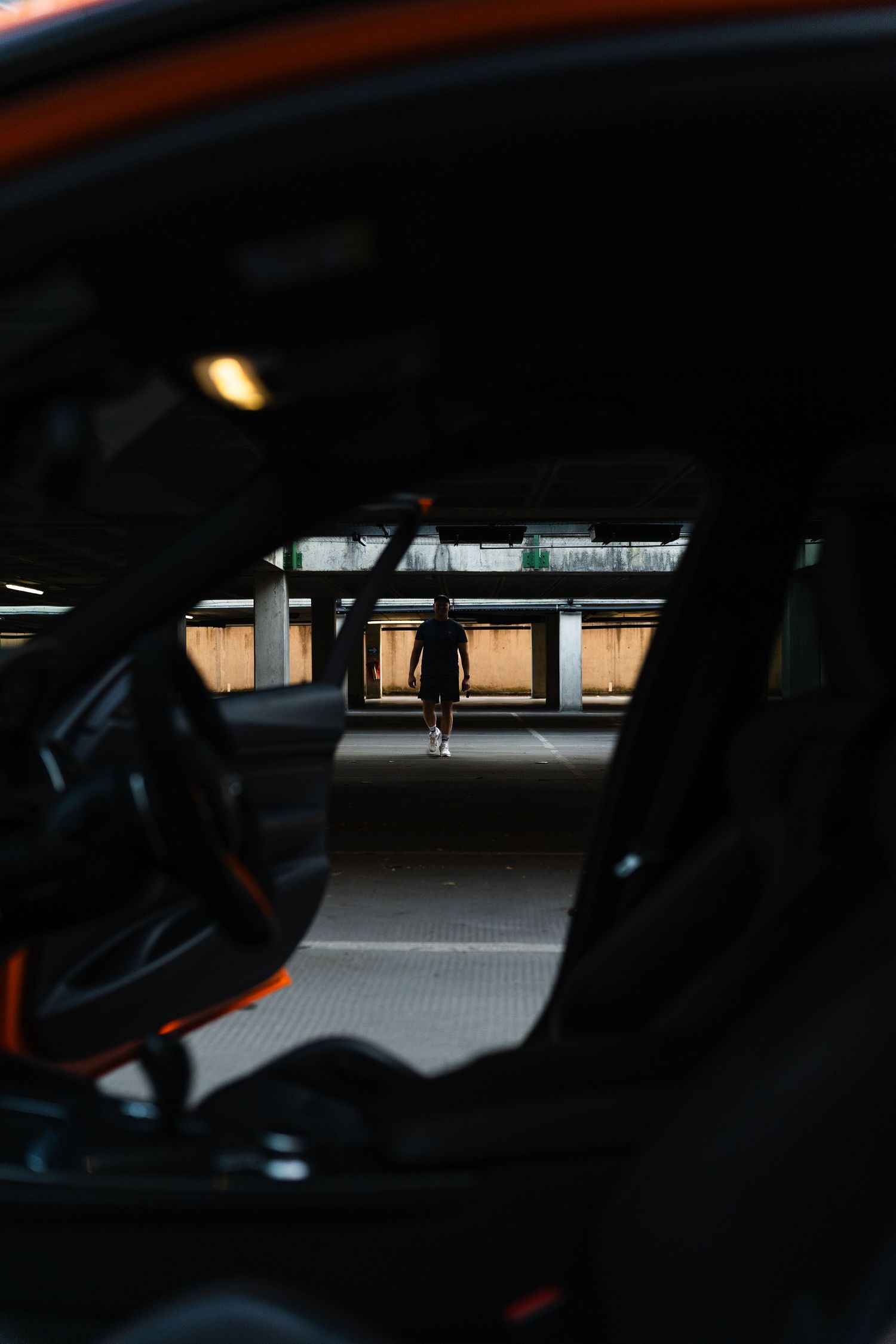 View through a dark sports car interior showing a silhouetted person standing in a dimly lit parking garage.