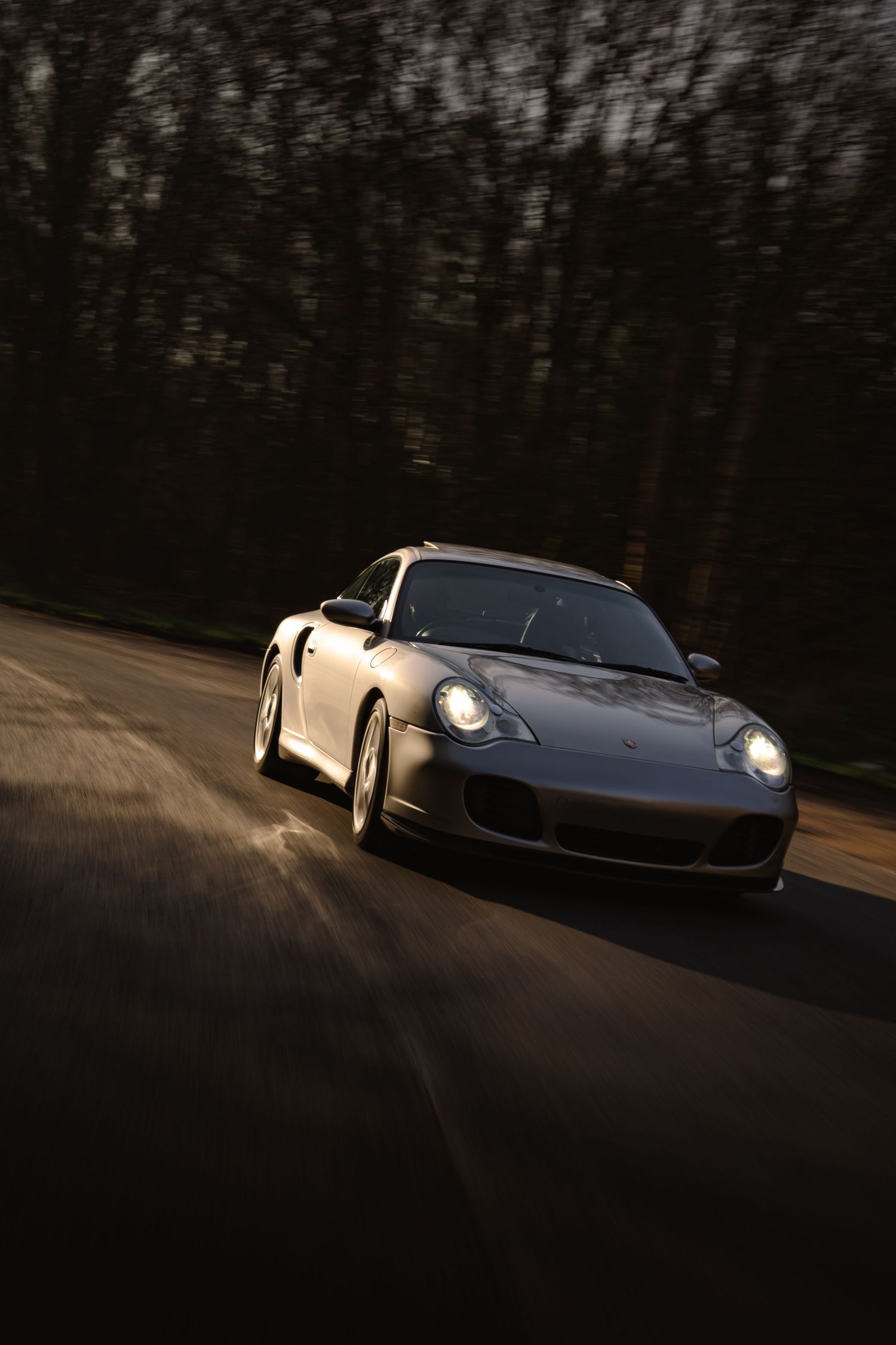 Silver Porsche 911 Turbo speeding along a dark tree-lined road with motion blur and headlights illuminated.