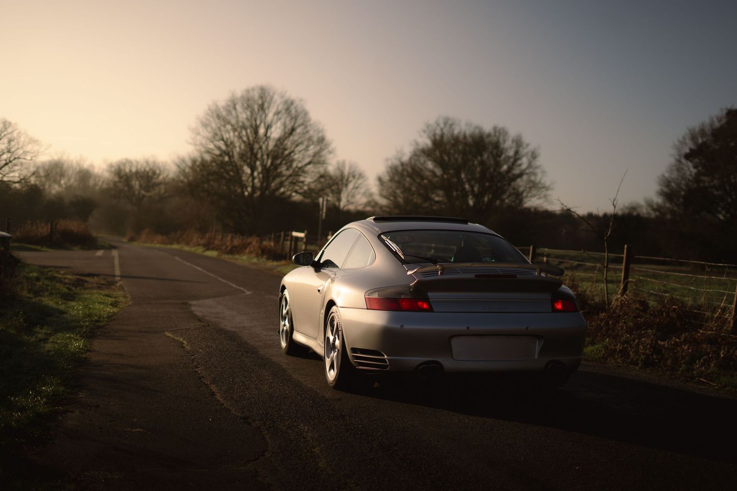Silver Porsche 911 rear three-quarter view on a country road bathed in warm golden sunset light.