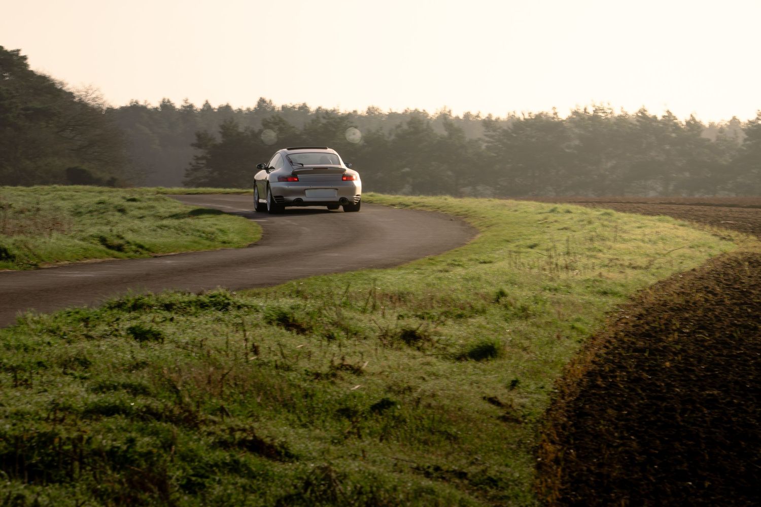 Silver Porsche 911 Turbo navigating a curved rural road surrounded by green fields at sunrise.