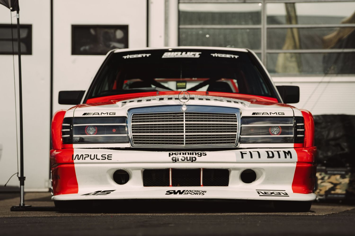 A Mercedes-Benz 190E DTM racing car in white and red livery with AMG branding, photographed head-on at a motorsport event.