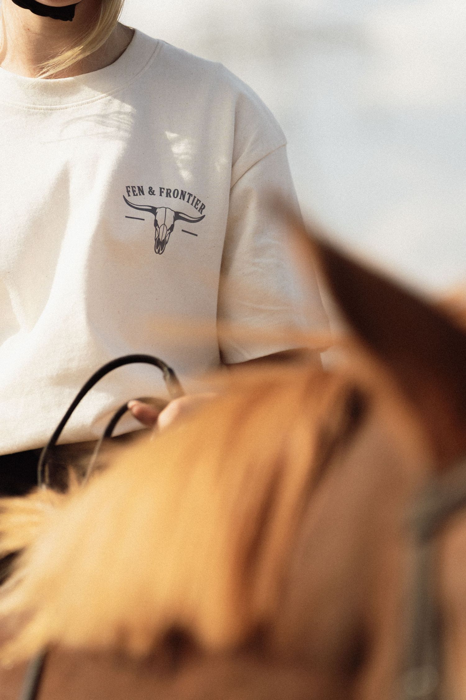Person wearing a cream Fen & Frontier graphic tee with bull skull logo, riding a brown horse outdoors.