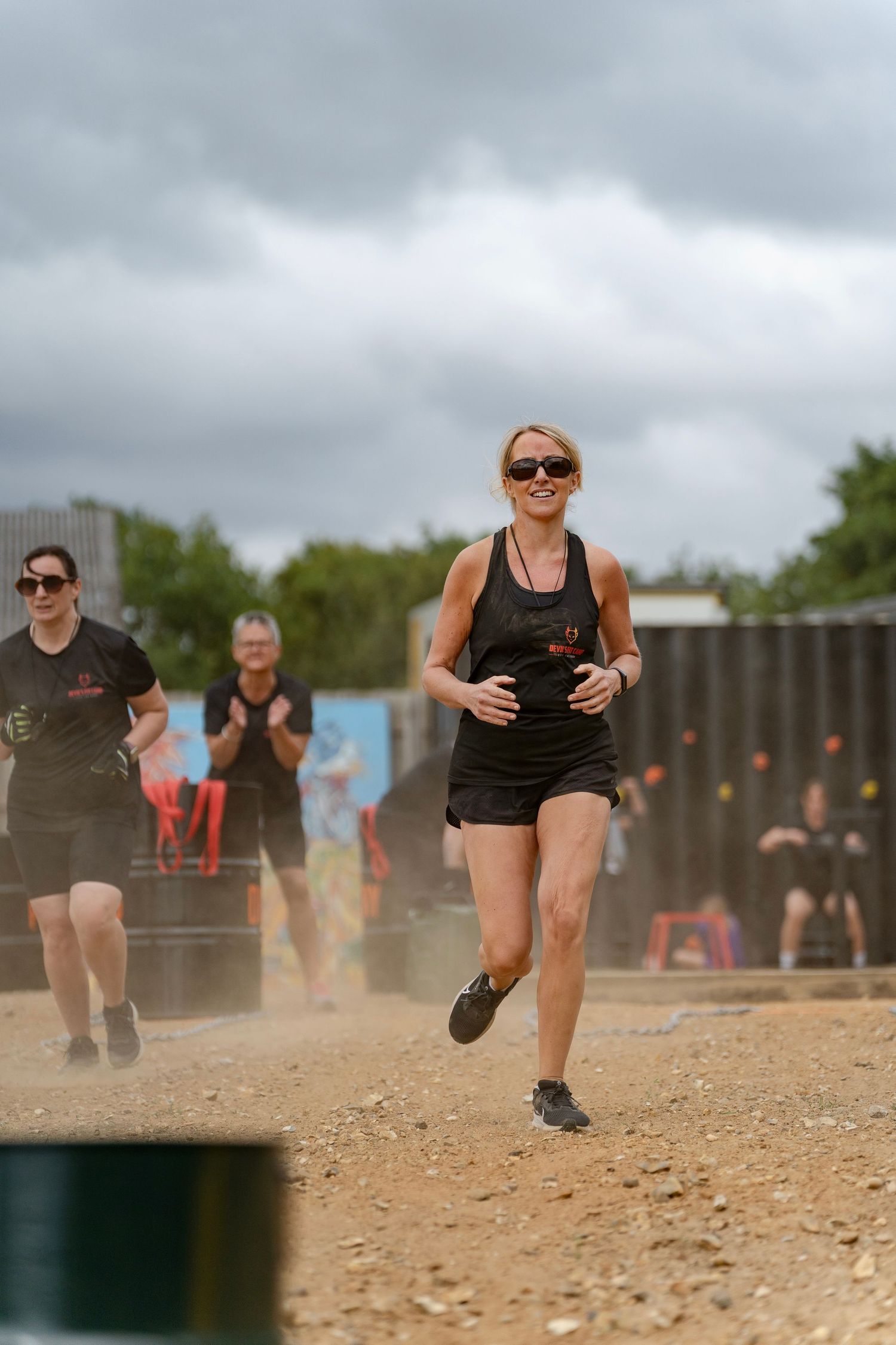 Woman in black athletic wear running confidently on a dirt track during an outdoor obstacle race event on a cloudy day.