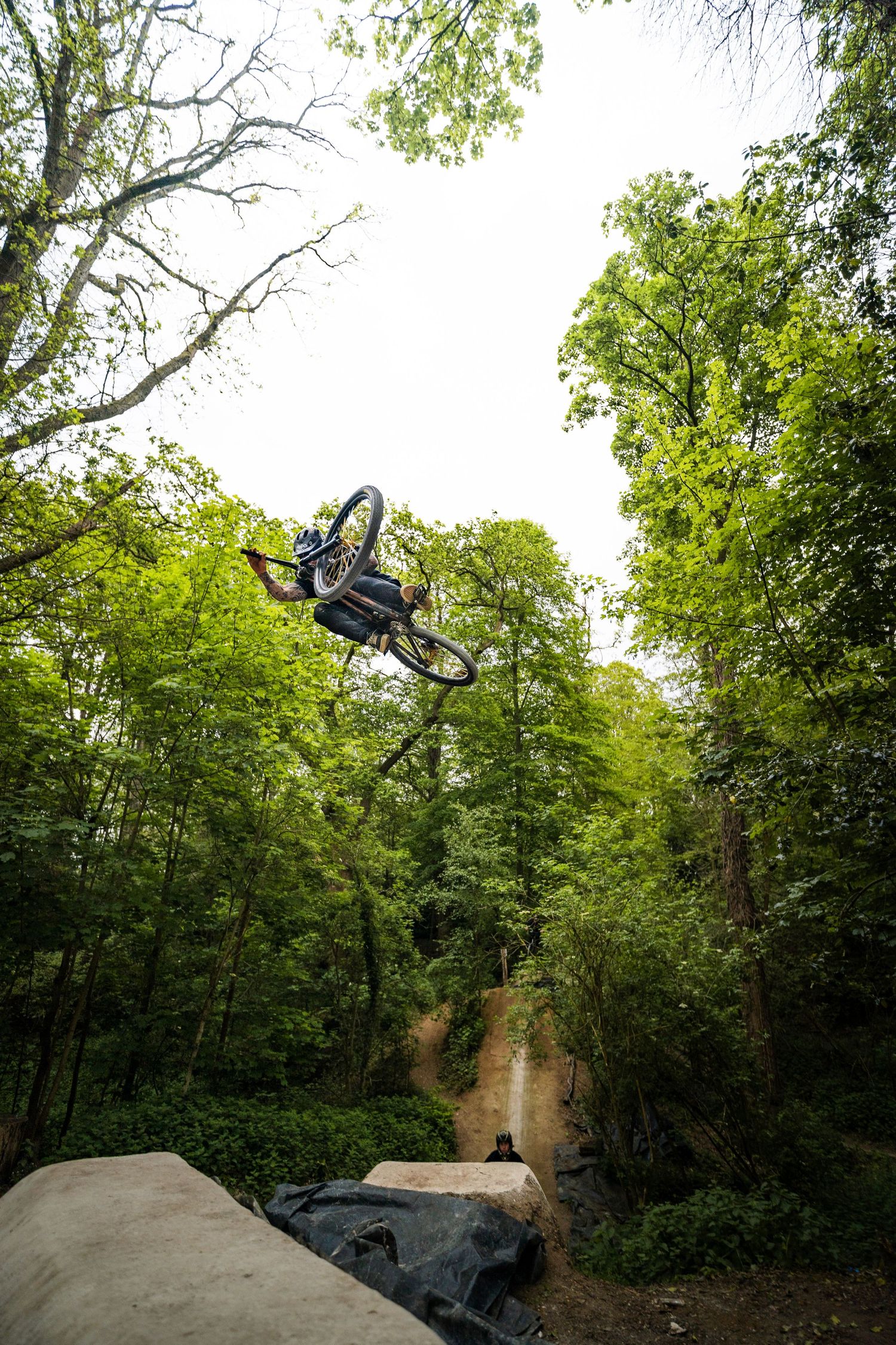 Mountain biker performing a high aerial trick jump over dirt ramps in a lush green forest trail.