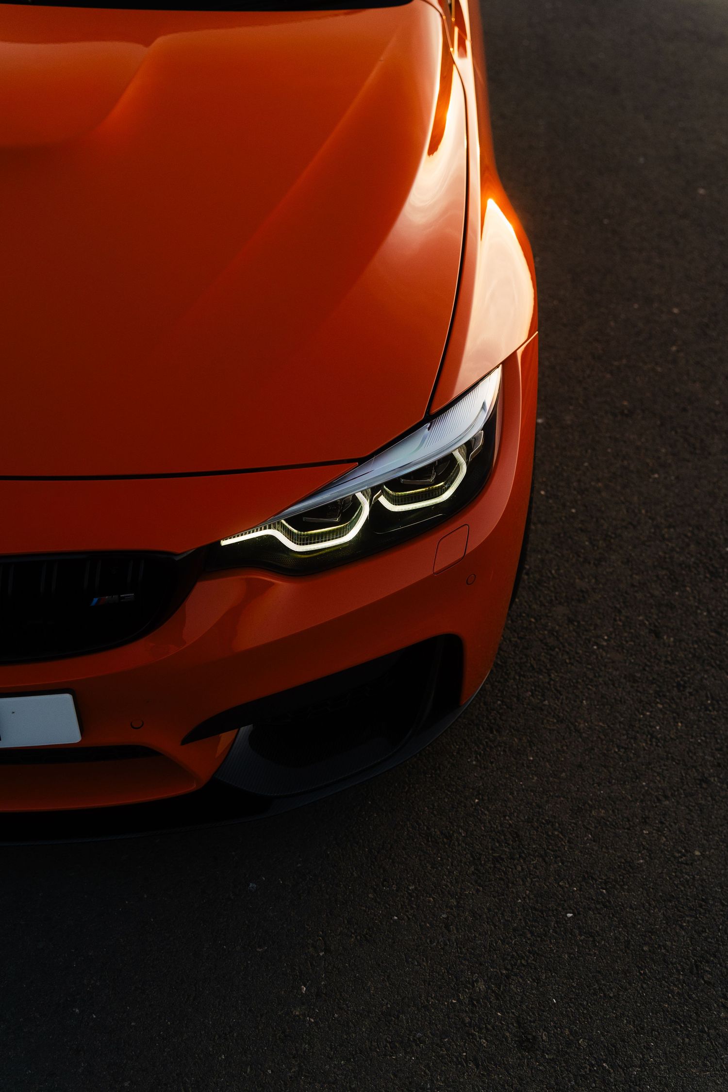 Vibrant orange BMW M4 sports car front quarter view with illuminated LED headlights on dark asphalt.