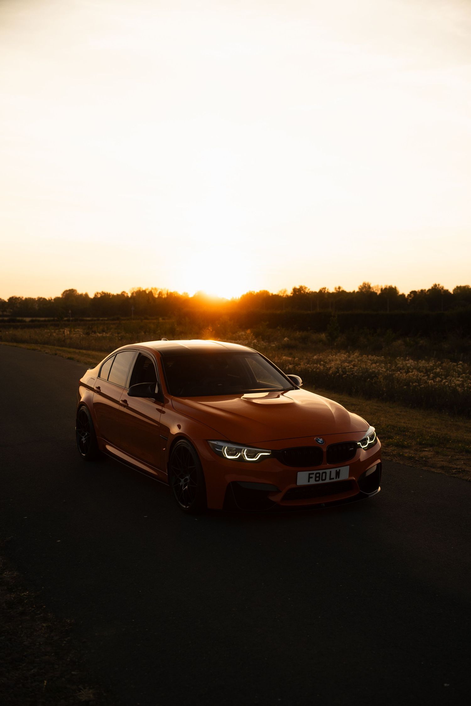 Orange BMW M3 F80 parked on a road at golden sunset with trees silhouetted in the background.