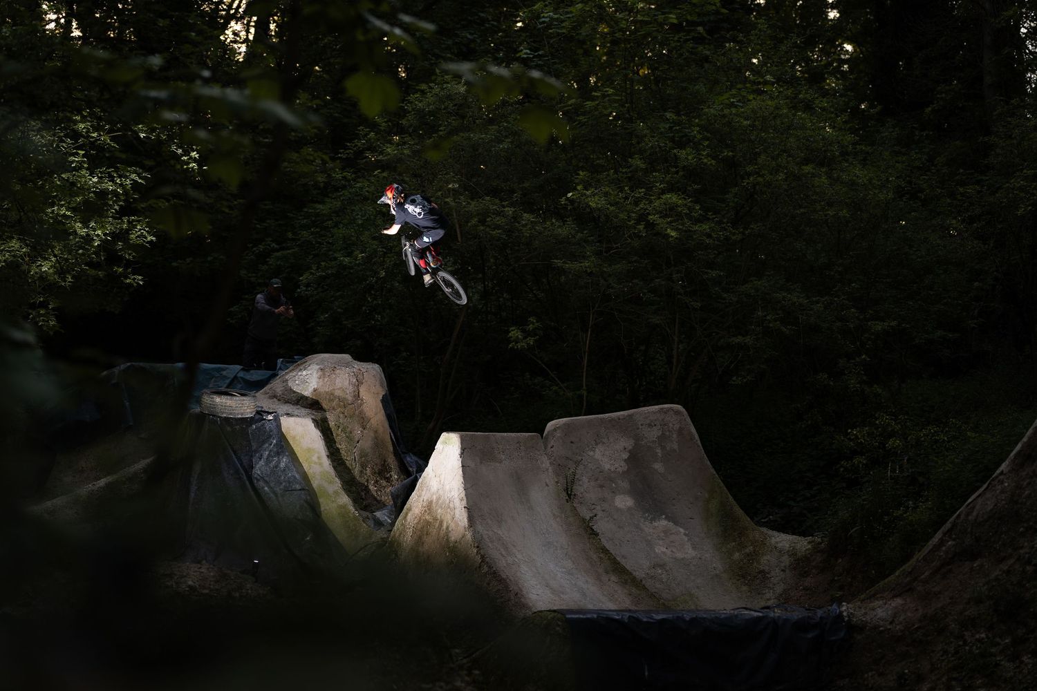A mountain biker performs a high jump trick over concrete ramps in a forested bike park at dusk.