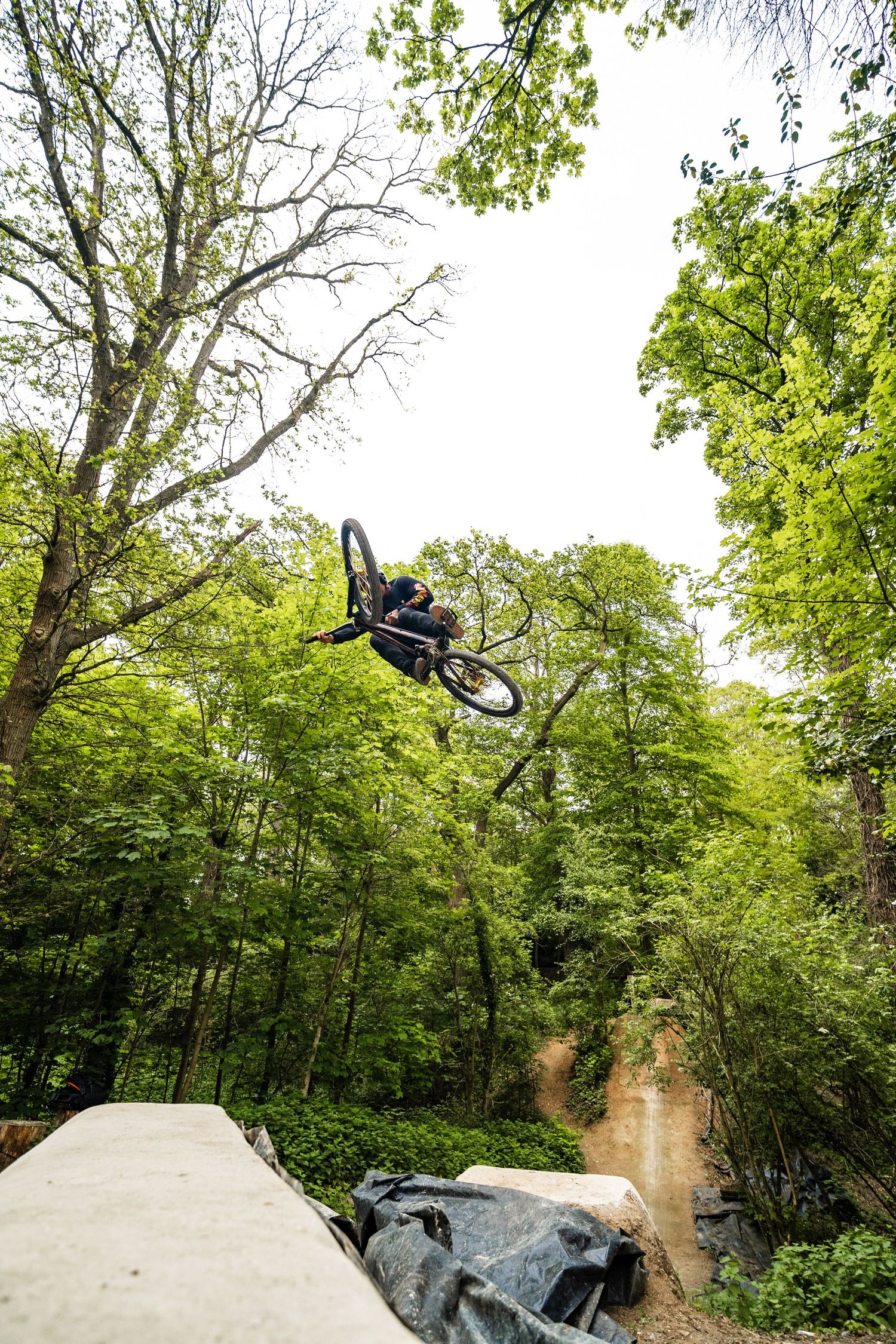 A mountain biker performs a high aerial jump trick over rocks in a lush green forest setting.