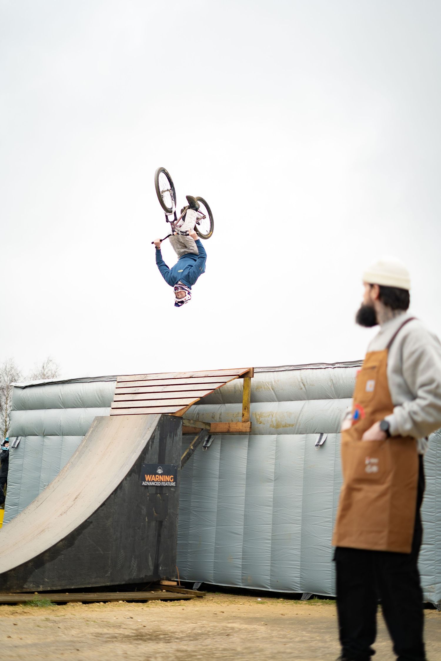 A BMX rider performs a high aerial trick while a bearded barista in a brown apron watches nearby.