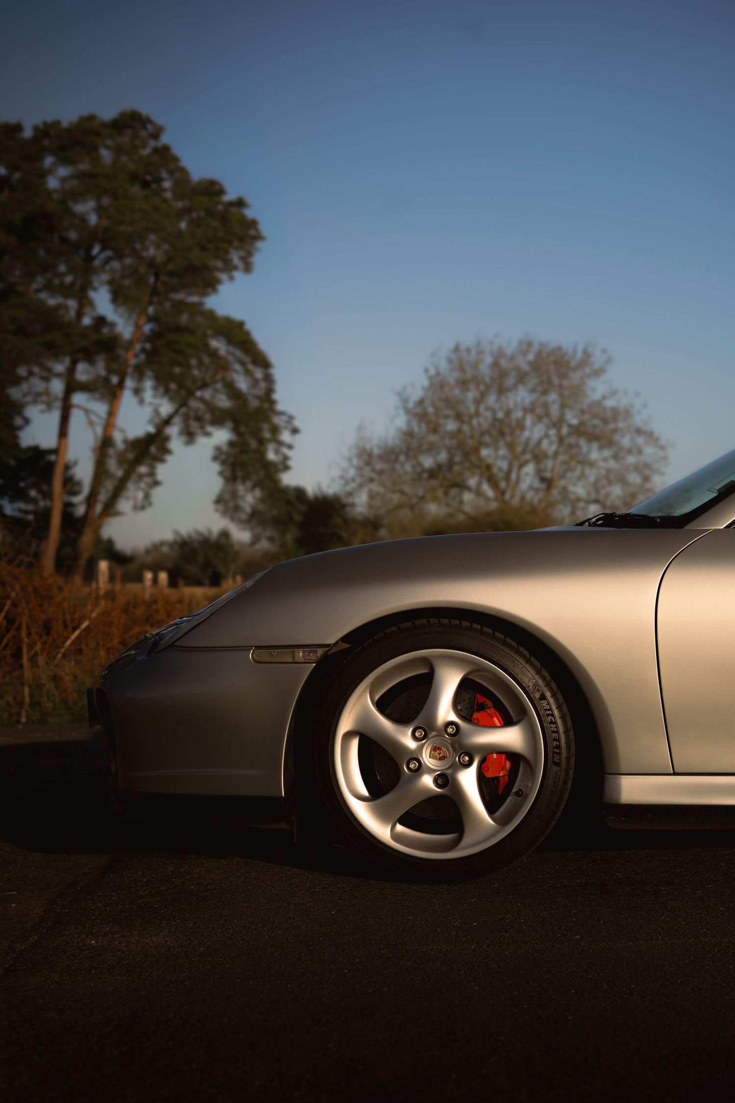 Side profile of silver Porsche 911 wheel with red brake calipers at golden hour in countryside.