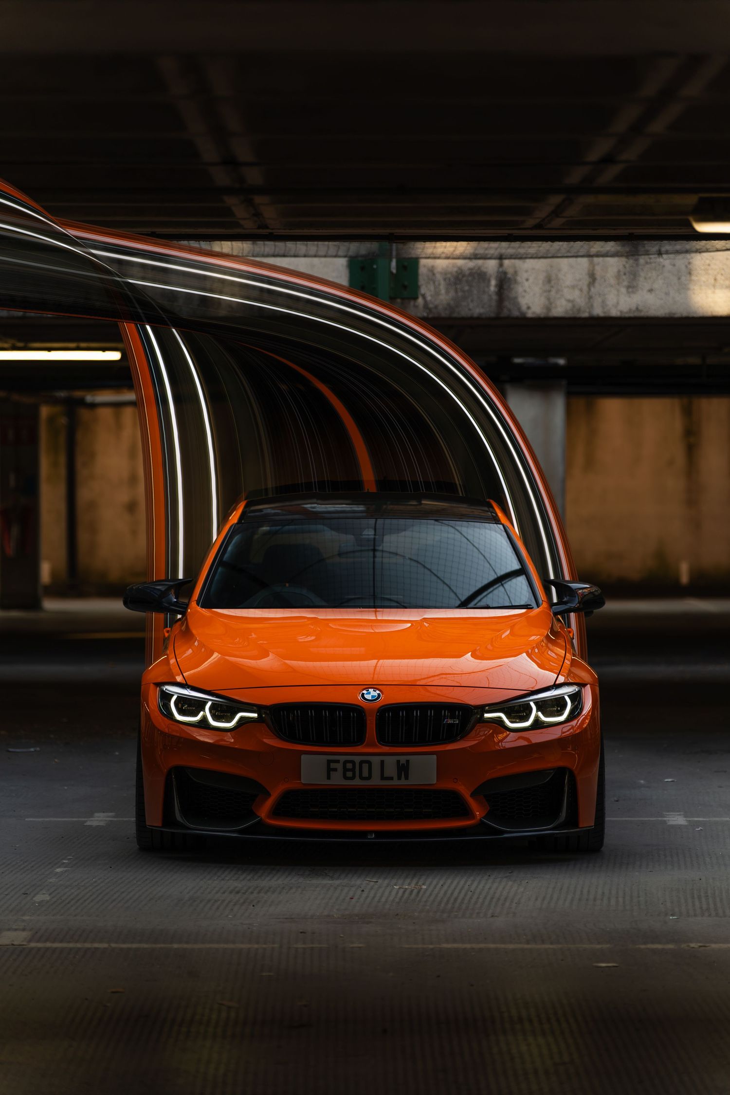 Orange BMW M4 parked in a dark underground garage with dramatic light streaks reflecting off its glossy exterior.