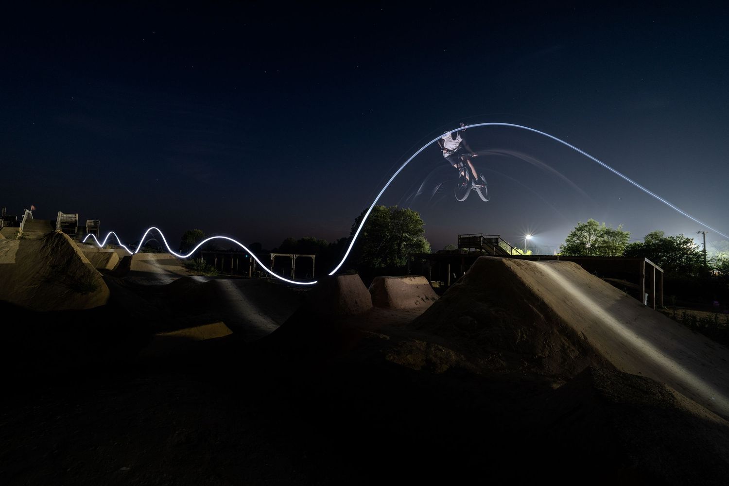 Long exposure night photo of a BMX rider performing tricks on a pump track, creating a glowing light trail against a dark sky.