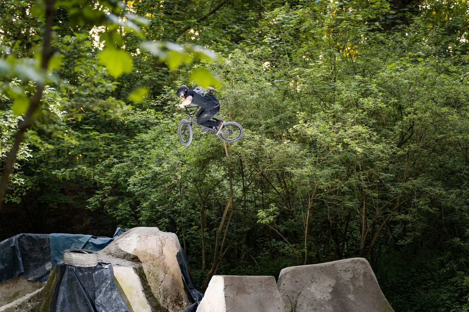 A BMX rider performs an aerial trick off a dirt jump ramp surrounded by lush green trees in an outdoor bike park.
