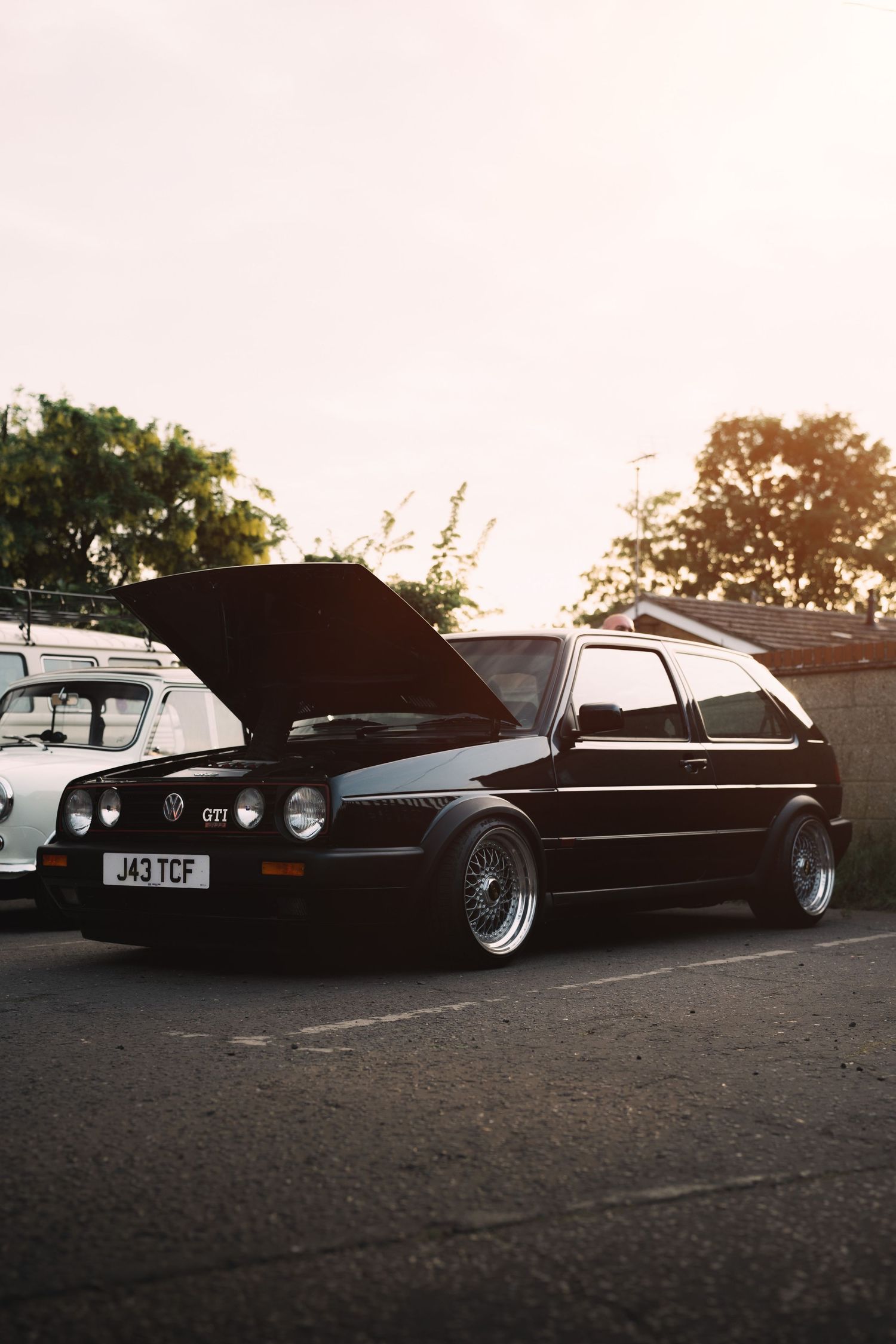 Black VW Golf GTI MK2 with hood open, lowered on BBS wheels, parked at a car meet during golden hour.