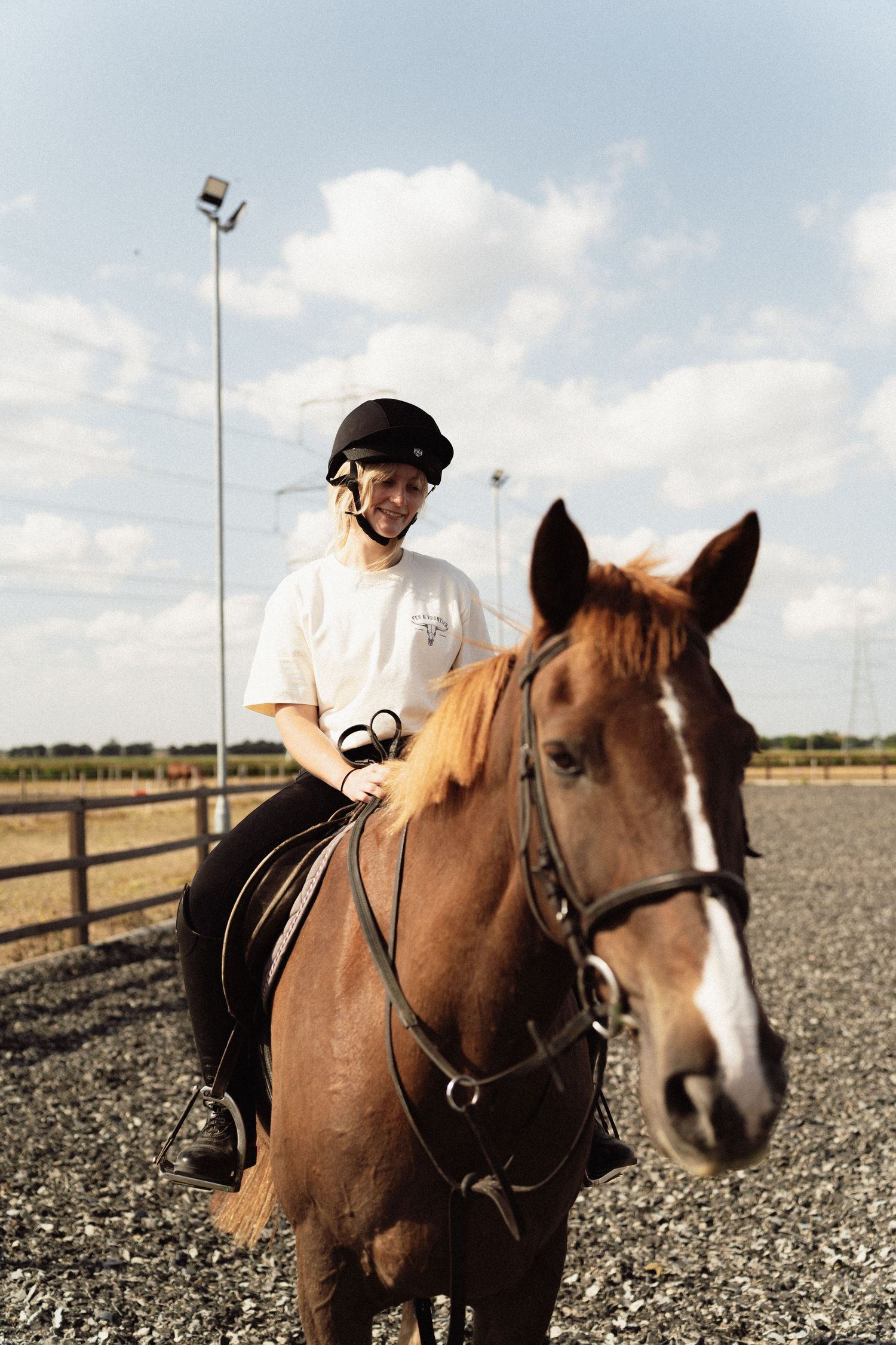 Smiling rider in black helmet on chestnut horse in outdoor arena under partly cloudy sky.
