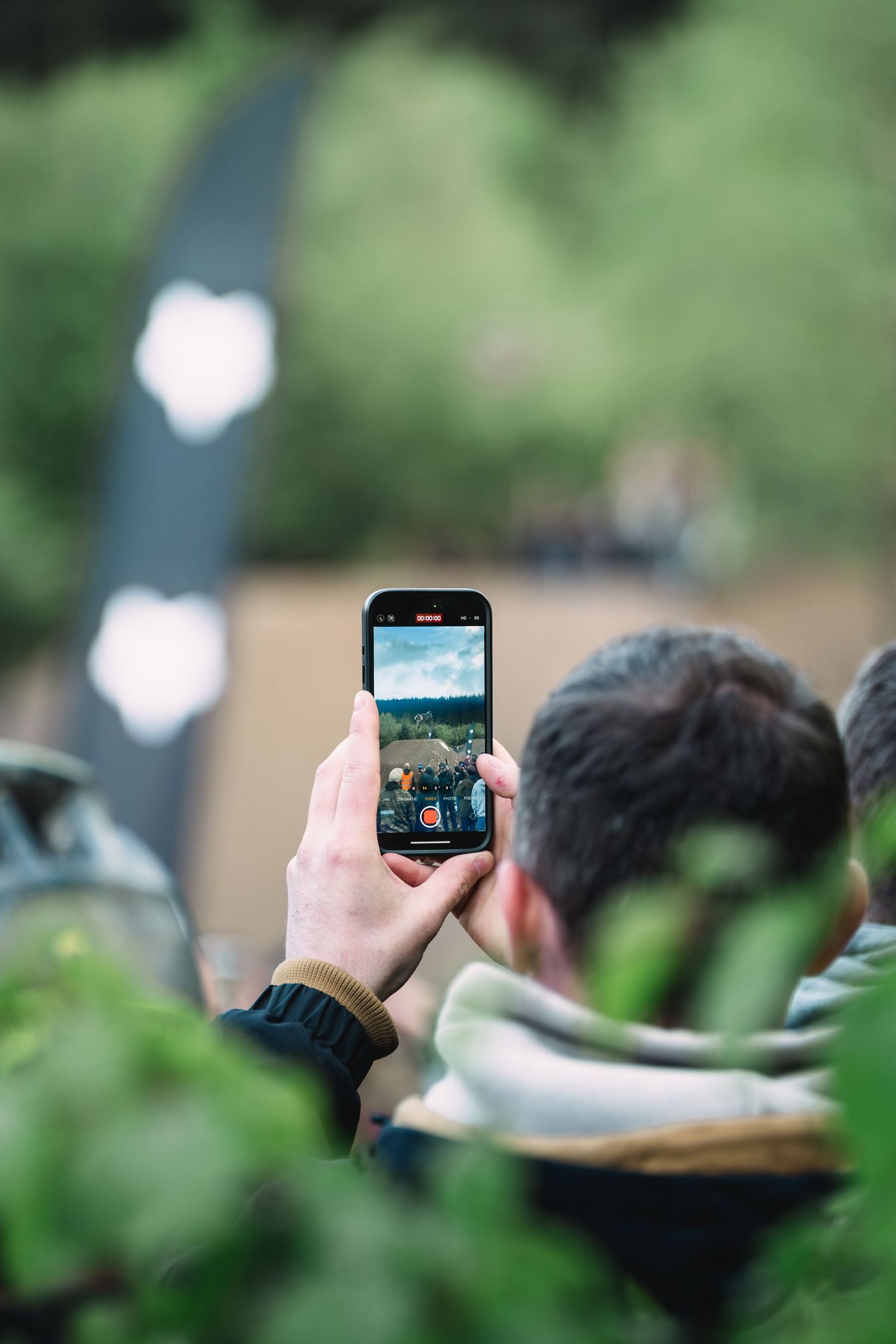 Man holding smartphone taking photo at outdoor event, captured from behind with green foliage in foreground.