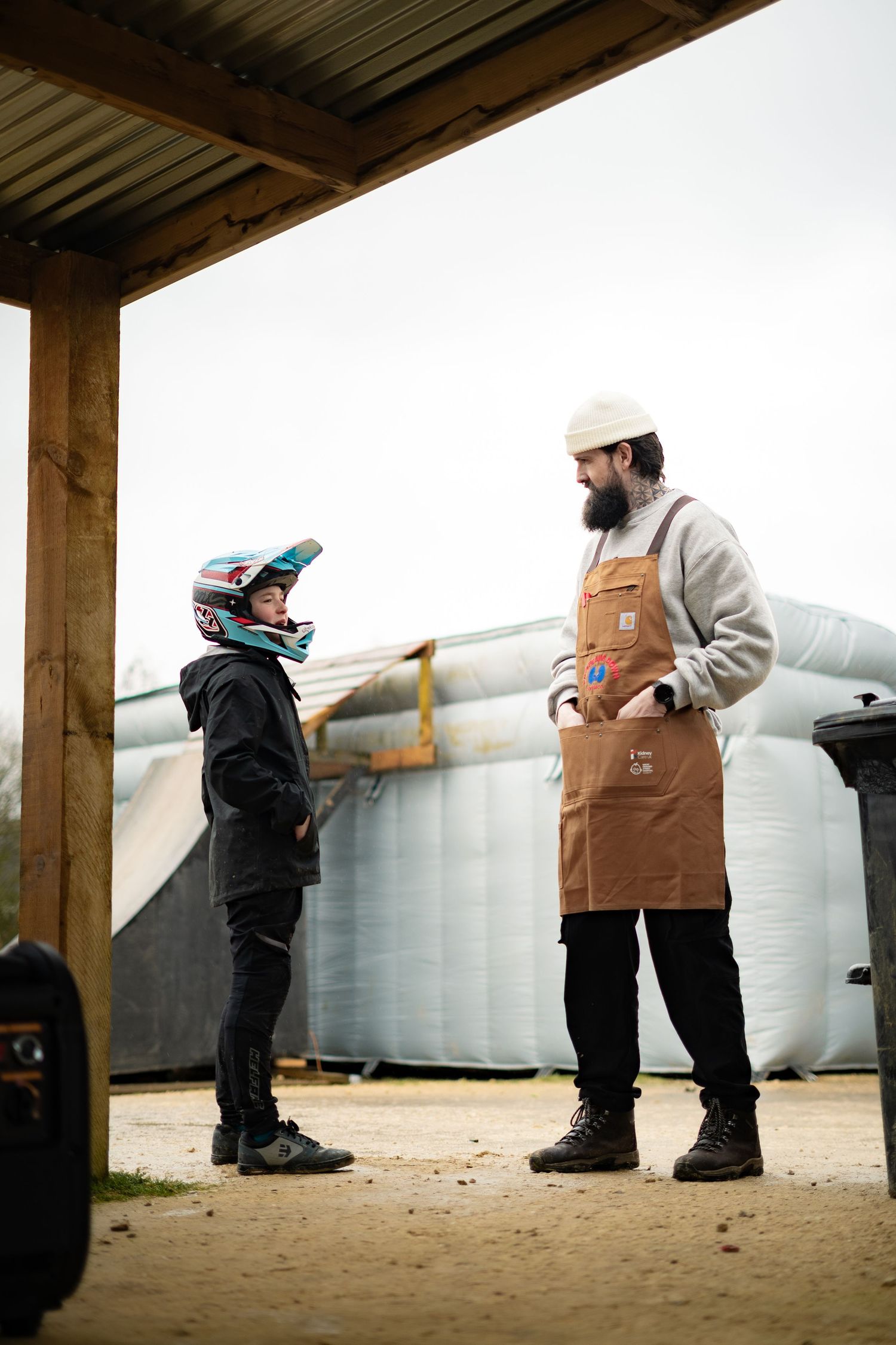 A bearded barista in a brown apron chats with a young BMX rider wearing a helmet outdoors.