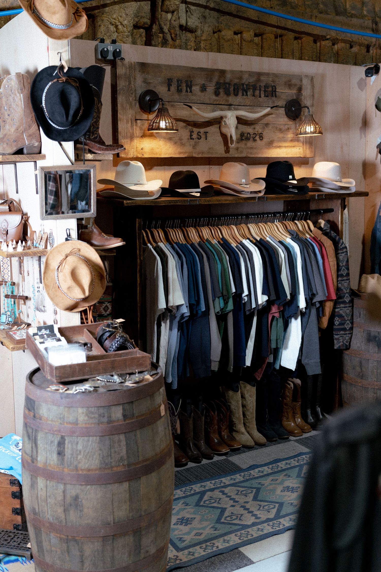 Rustic western boutique with cowboy hats, boots, and clothing rack displayed beside a wooden barrel.