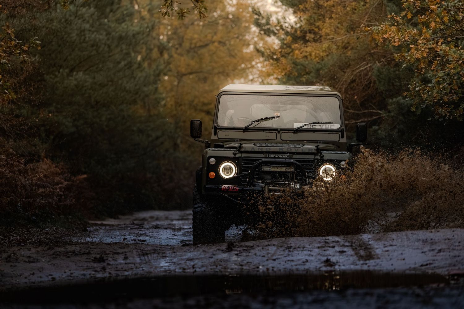 Classic Land Rover Defender driving through muddy puddle on a forest trail.