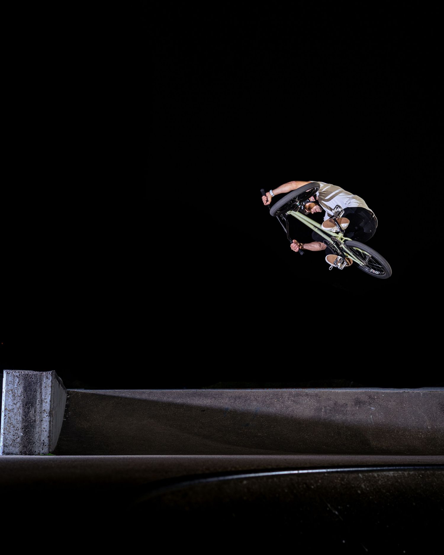 BMX rider performs a dramatic aerial trick over a dark concrete skate ramp at night.