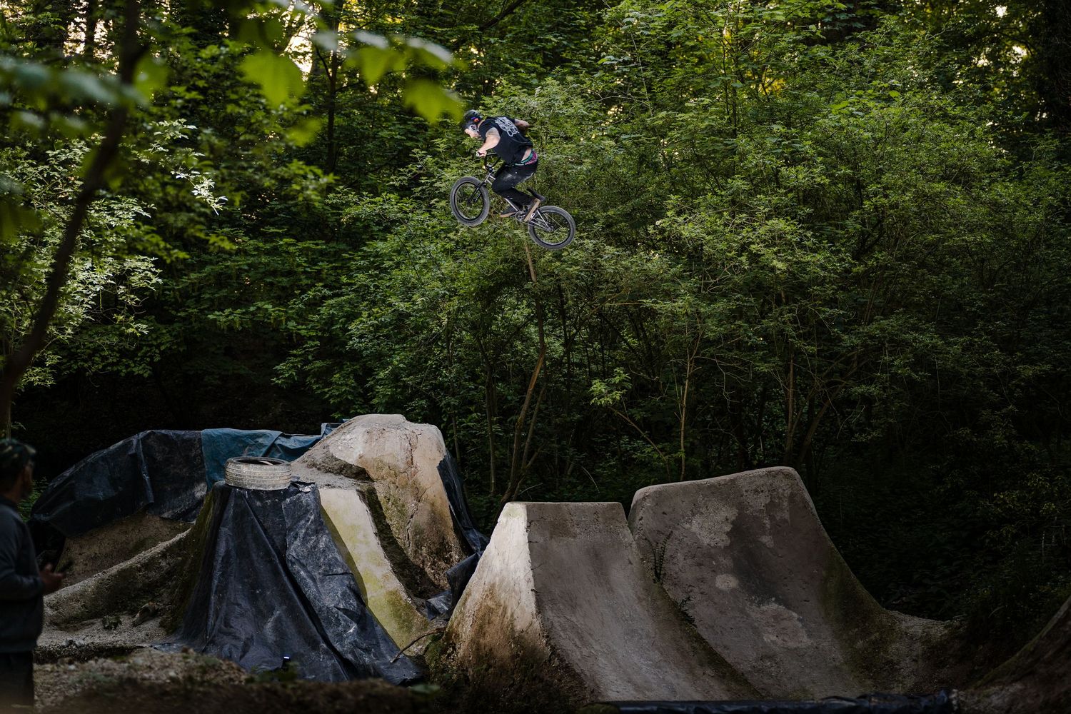 A mountain biker performs a high jump trick over concrete ramps in a forest bike park surrounded by lush green trees.