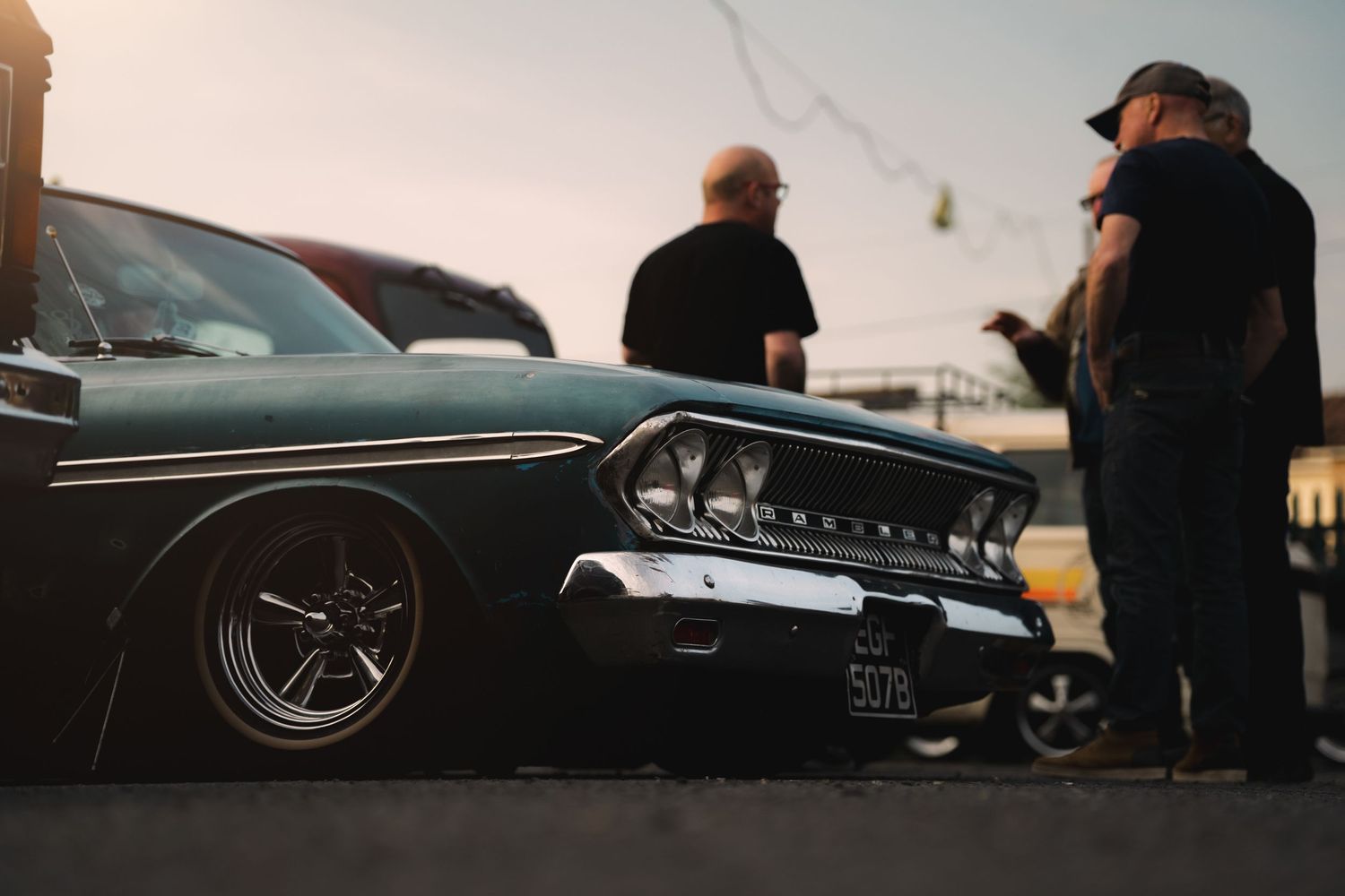 Lowered classic muscle car at a car show with two men standing nearby at sunset.