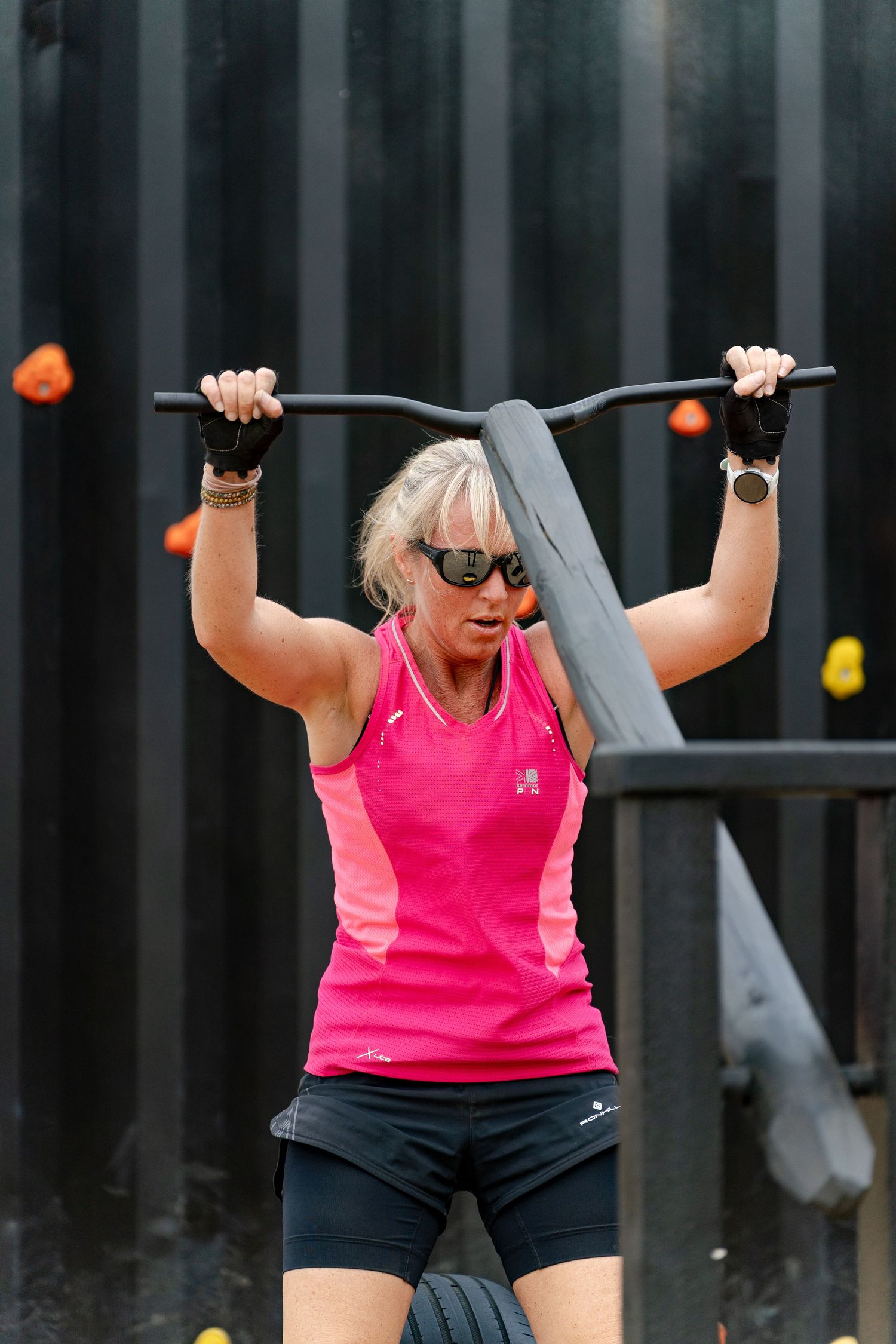 Woman in pink tank top doing pull-ups on outdoor gym bar, wearing sunglasses and workout gloves.