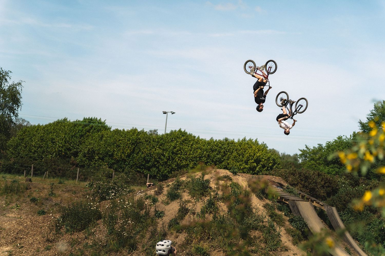 Two BMX riders perform backflip tricks simultaneously off a dirt jump ramp under a clear blue sky.