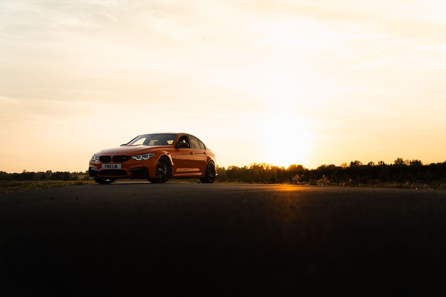 A golden BMW sedan parked on an open road during a stunning sunset with warm orange hues filling the sky.
