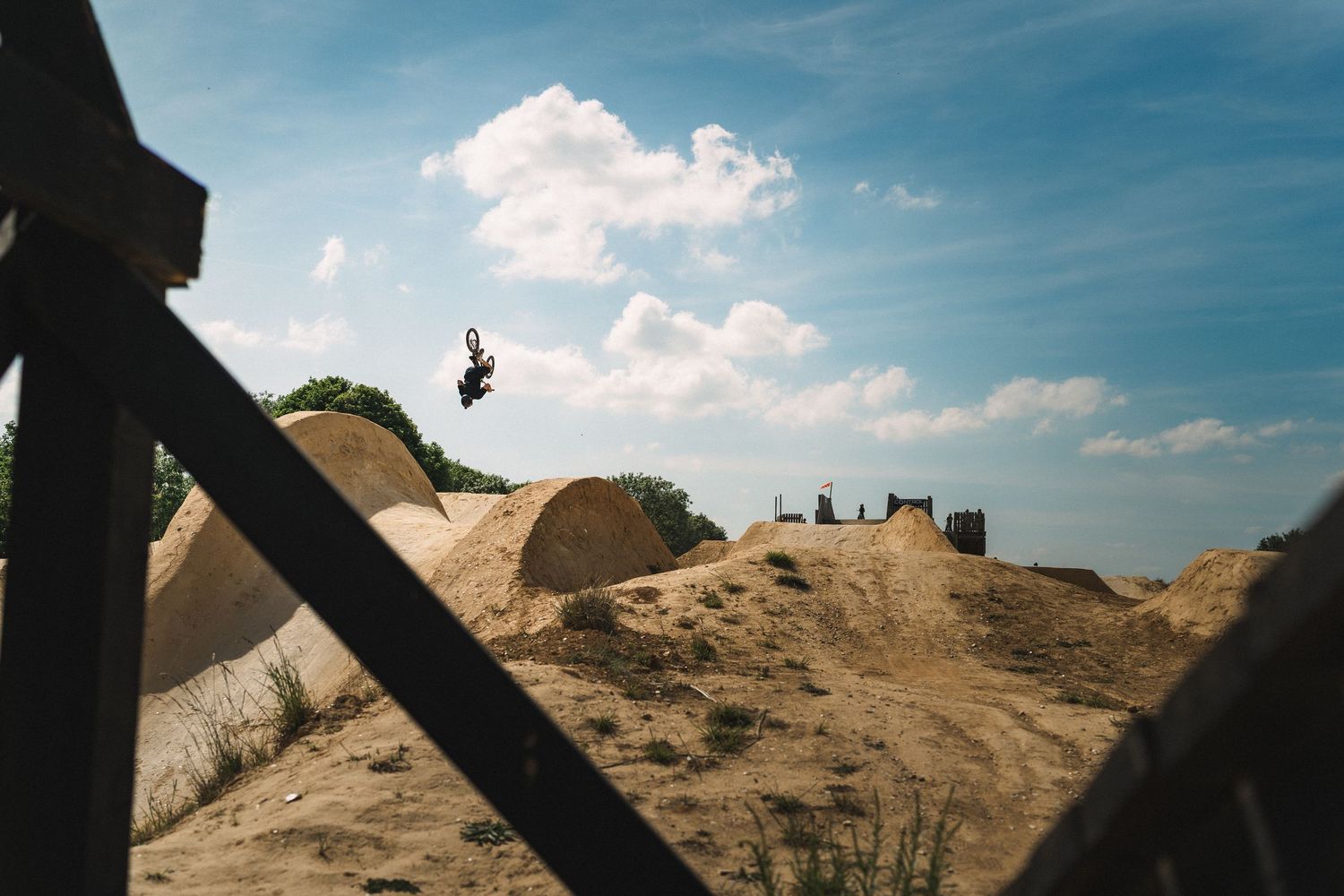 A person performs a high aerial trick over sandy dirt jumps at an outdoor bike park under a partly cloudy sky.