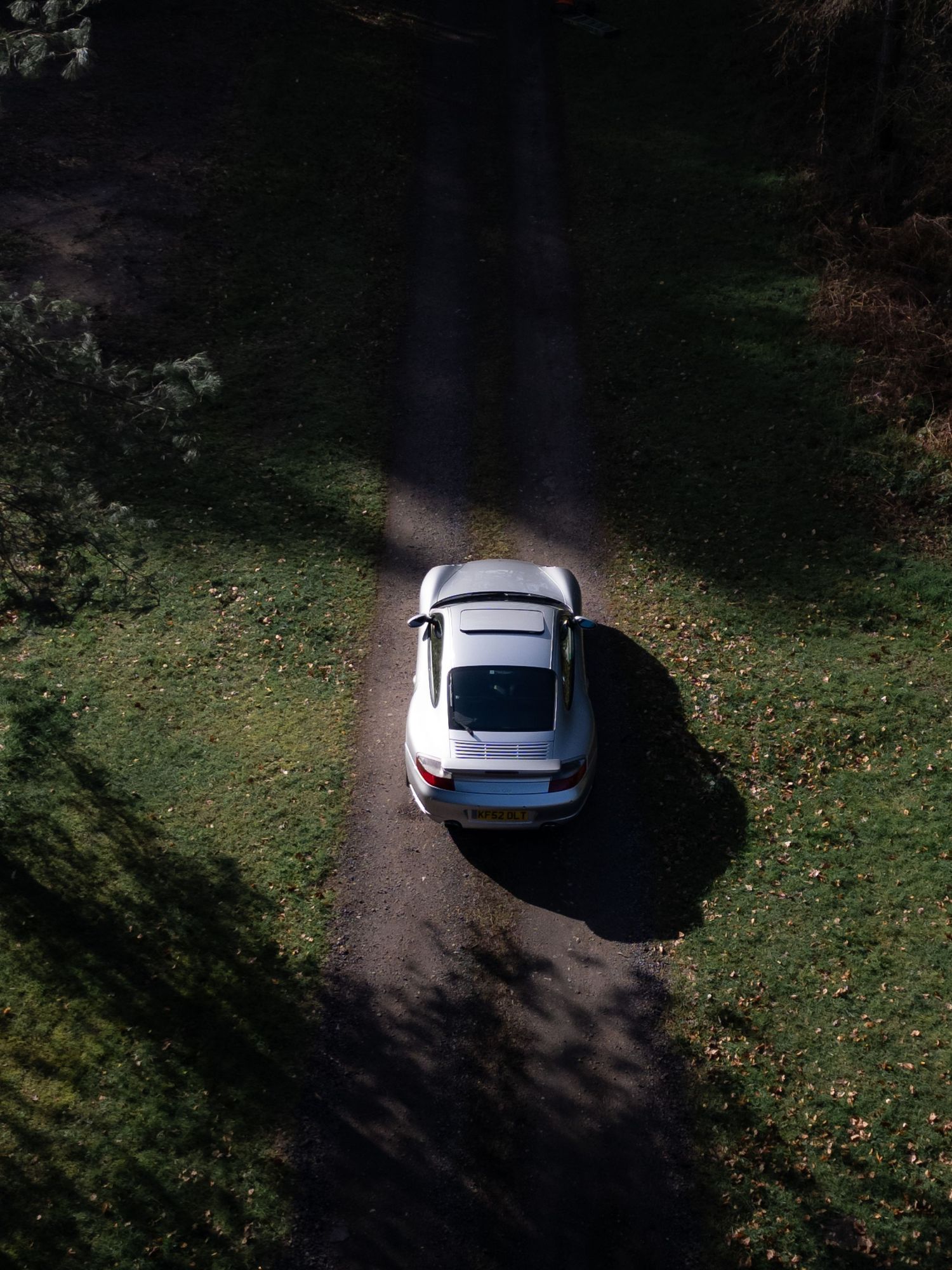 Aerial rear view of a silver Porsche 911 GT3 on a leaf-covered forest path in bright autumn sunlight.