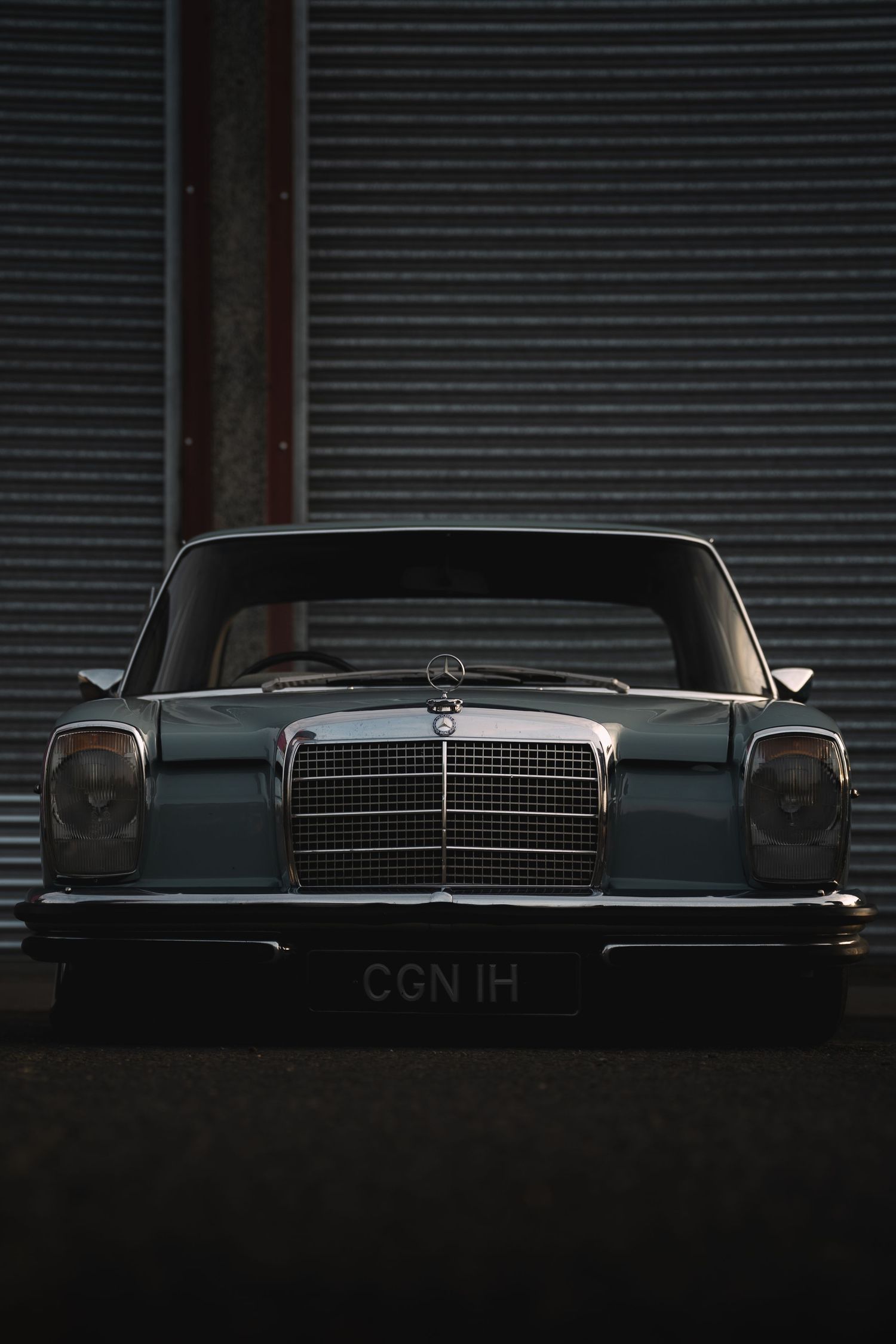 Vintage Mercedes-Benz classic car with chrome grille, dark teal finish, license plate CGN 1H, parked against corrugated metal wall.