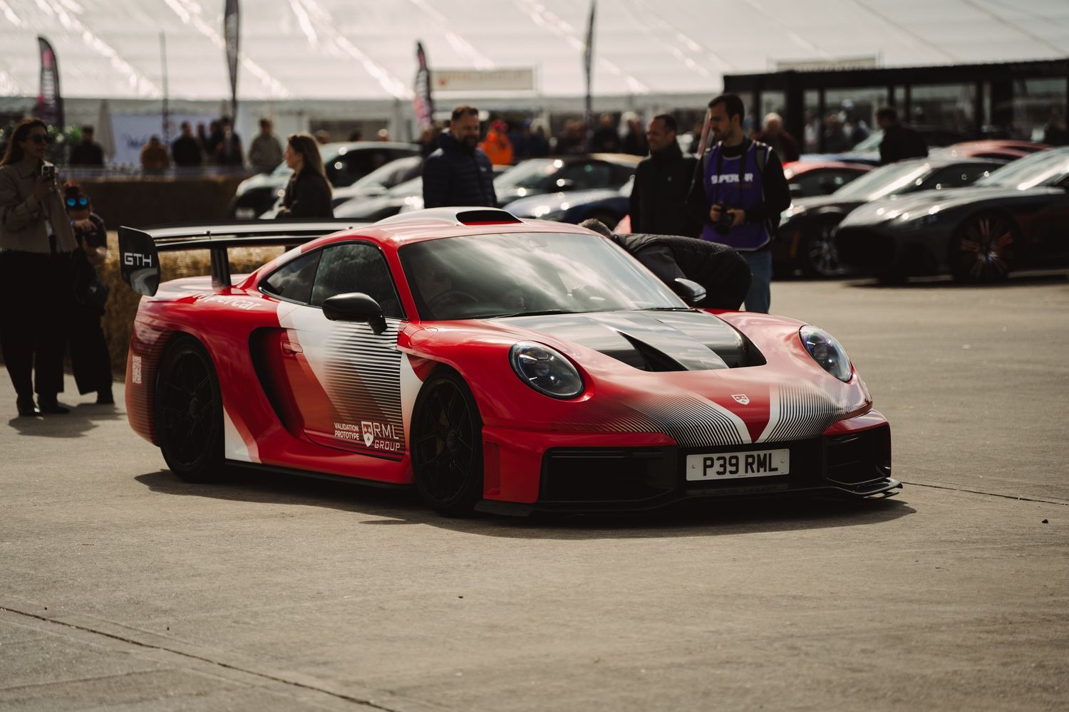 Red and white Porsche GT racing car with livery on track at motorsport event with spectators in background.