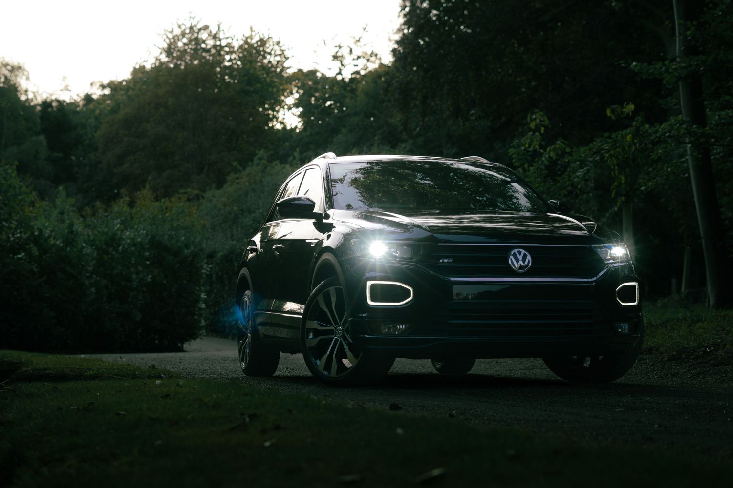 Black Volkswagen T-Roc SUV with glowing LED headlights parked among lush green trees at dusk.