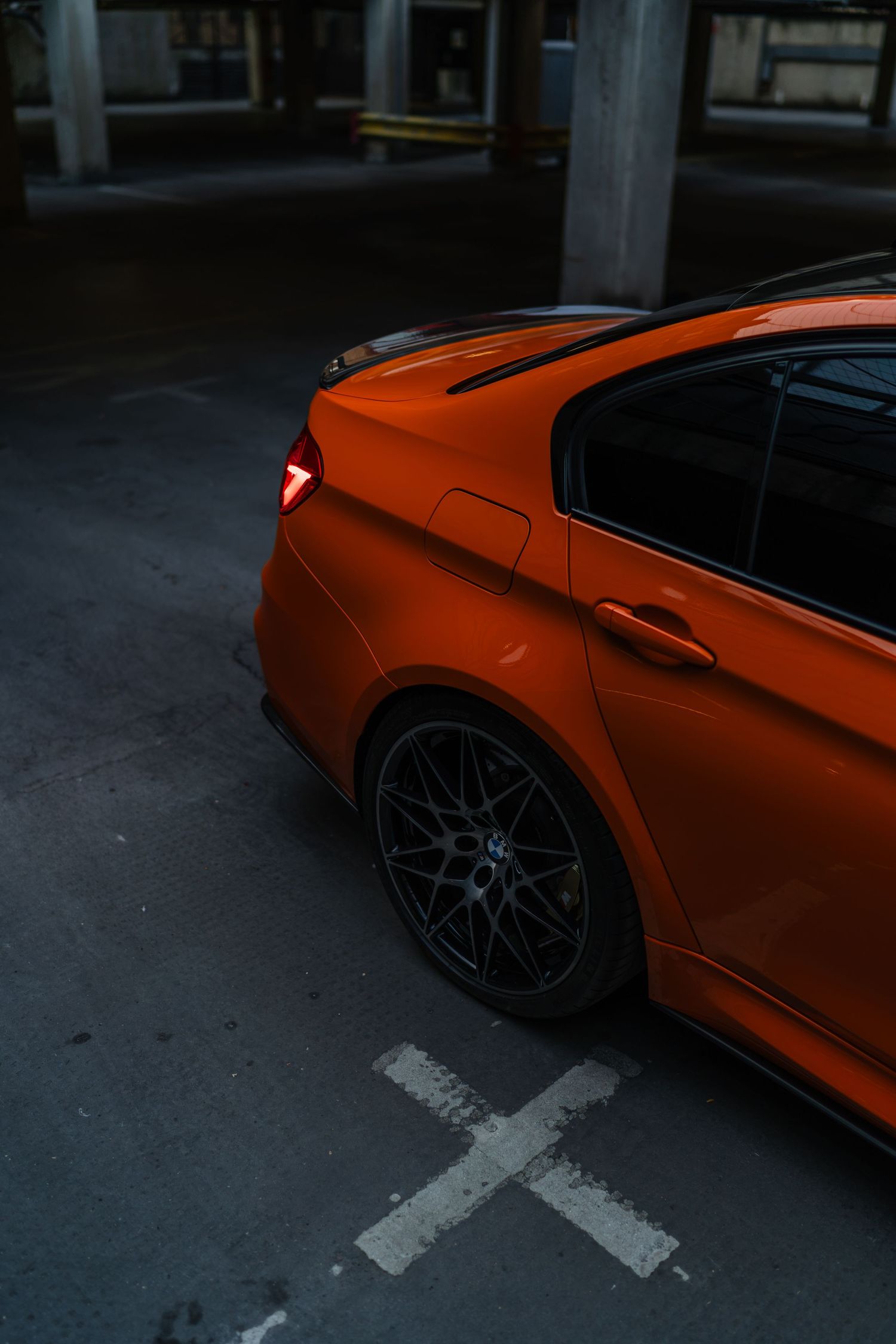 Vibrant orange BMW M3 sedan parked in a dark garage, showcasing sporky rear spoiler and black alloy wheels.