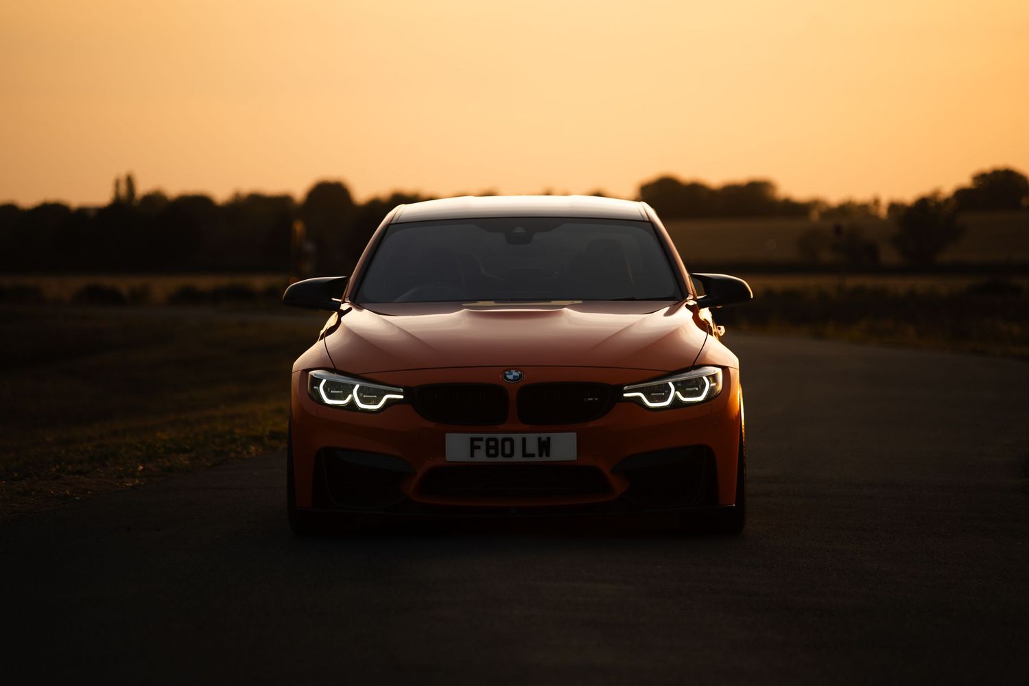 Orange BMW M4 captured from the front at golden hour sunset on an empty road with warm glowing sky backdrop.