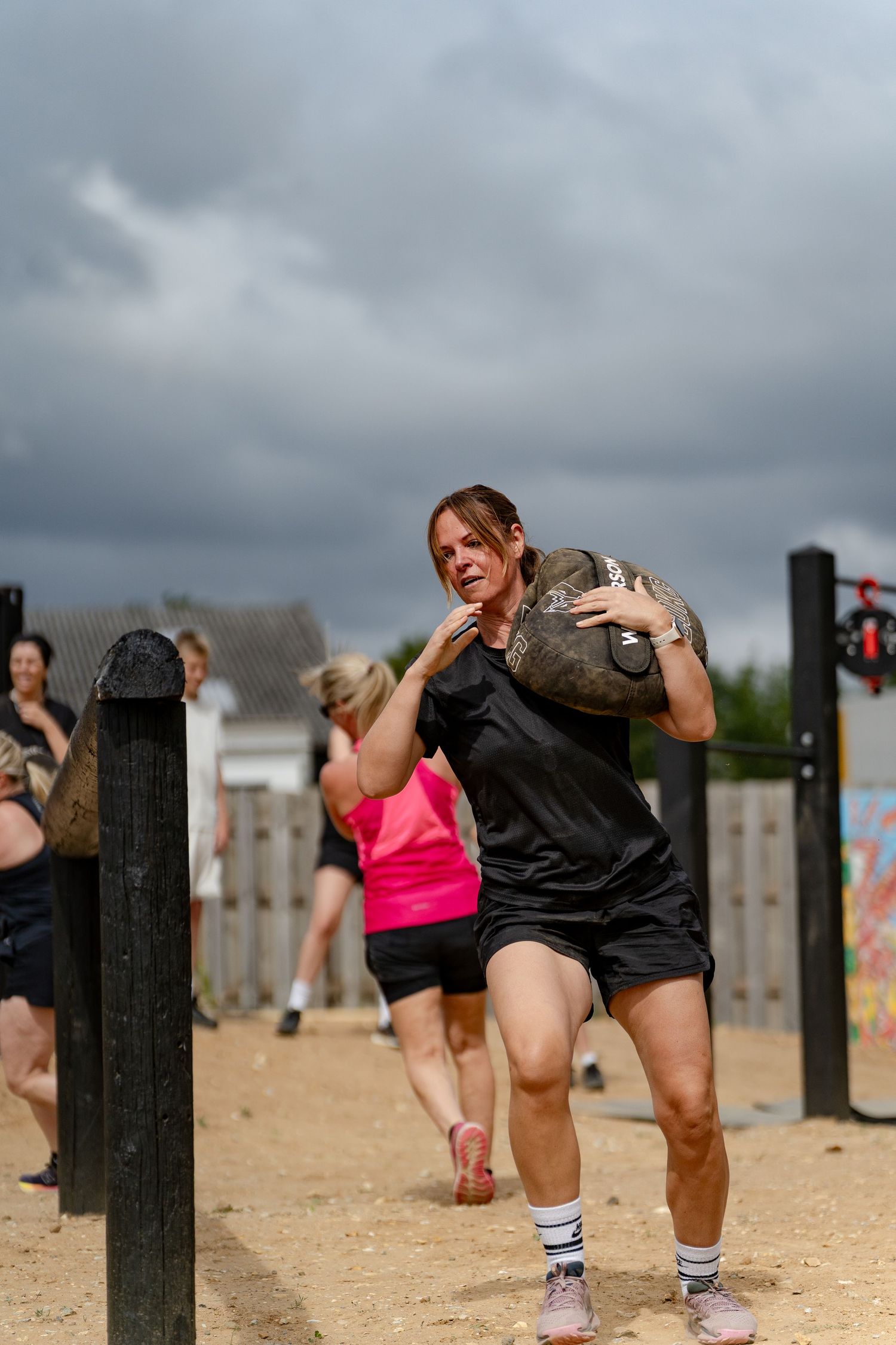 Woman carries heavy sandbag on shoulder during outdoor fitness challenge on sandy ground with stormy sky backdrop.
