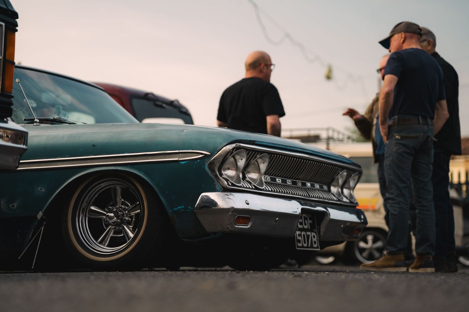 Vintage teal lowrider classic car at an outdoor car show with two men admiring it in the background.