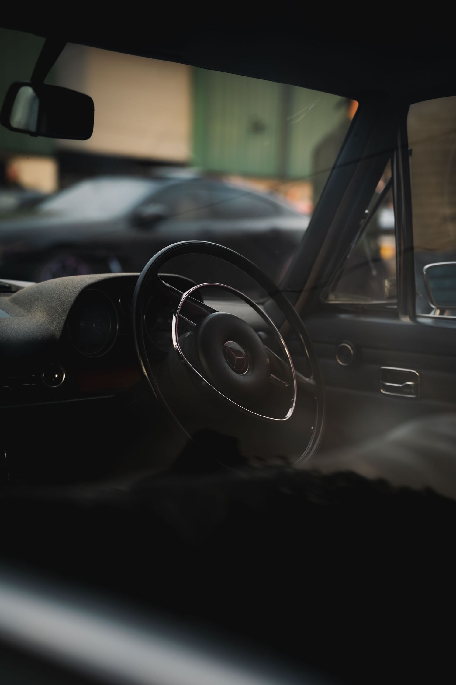 Dark moody interior view of a Mercedes-Benz steering wheel with blurred city background through windshield.