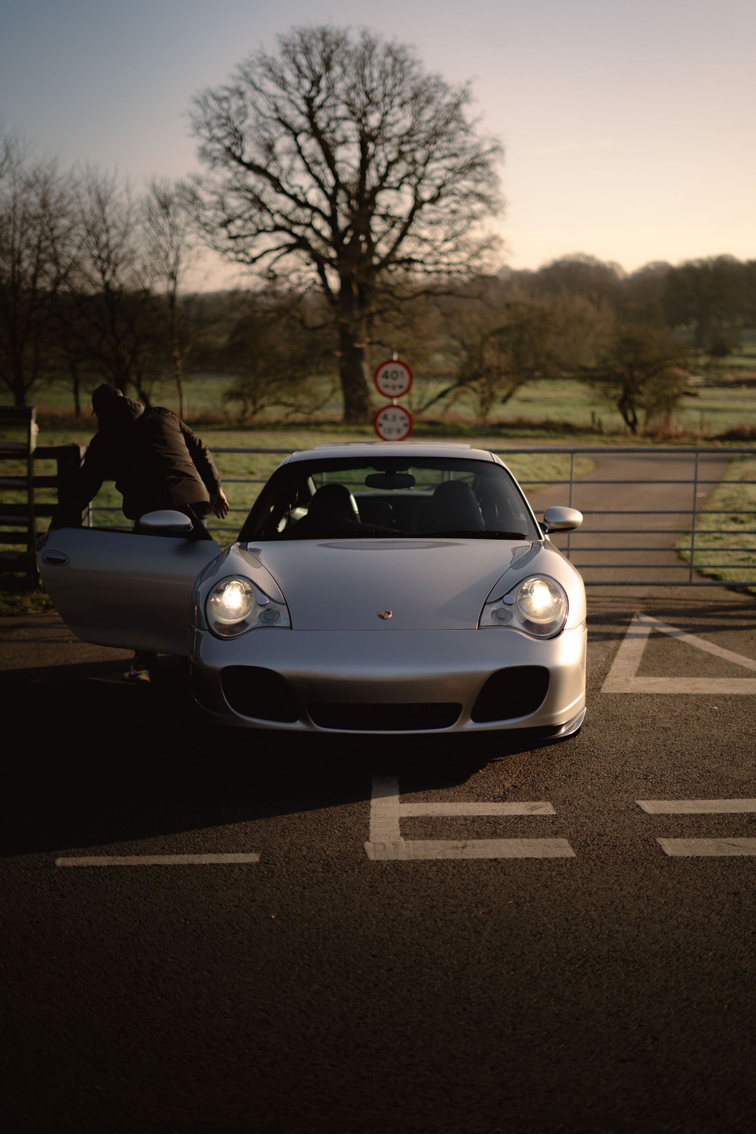 Front view of a silver Porsche 911 with headlights on in a car park at dusk with a person nearby.