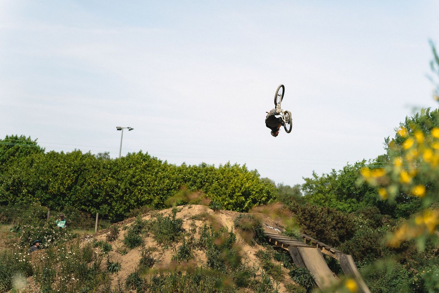 BMX rider performs a dramatic aerial trick high above a dirt jump park surrounded by lush green bushes and trees.