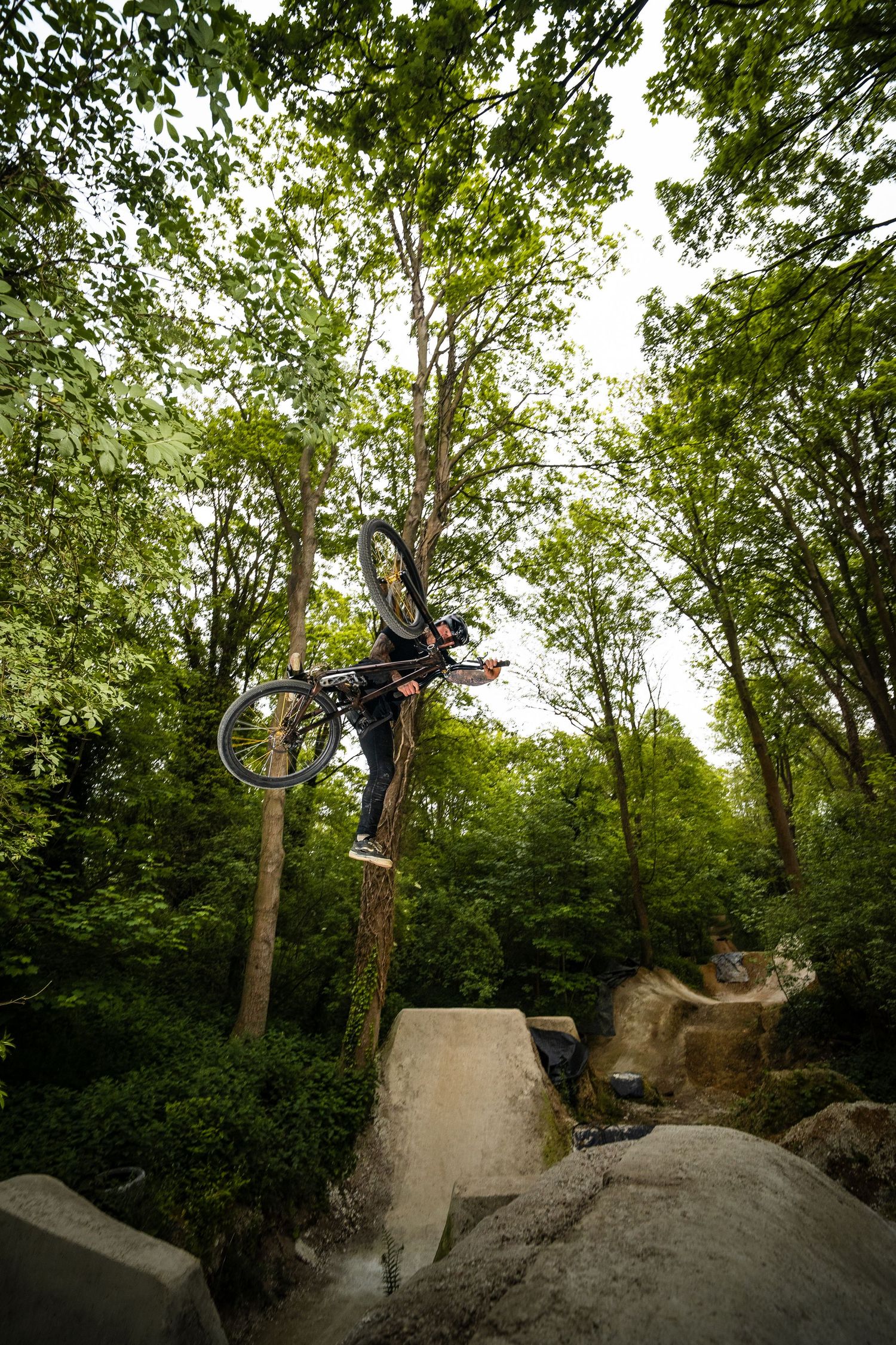BMX rider performing a high aerial trick over concrete blocks in a lush green forest setting.