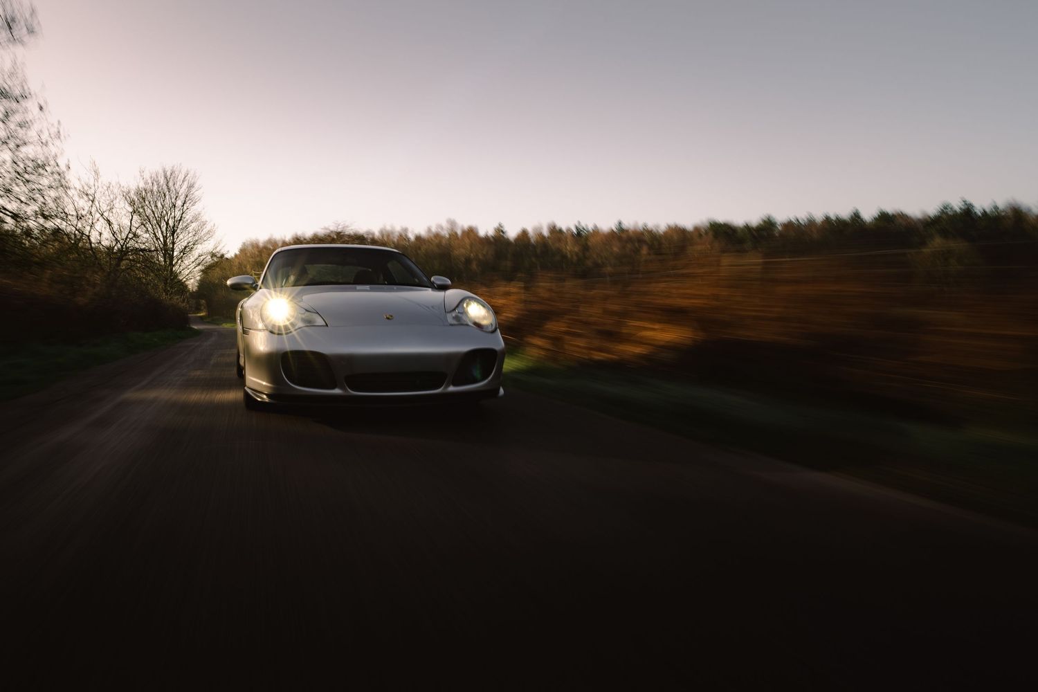 Silver Porsche 911 captured head-on driving on a rural road during golden hour sunset.