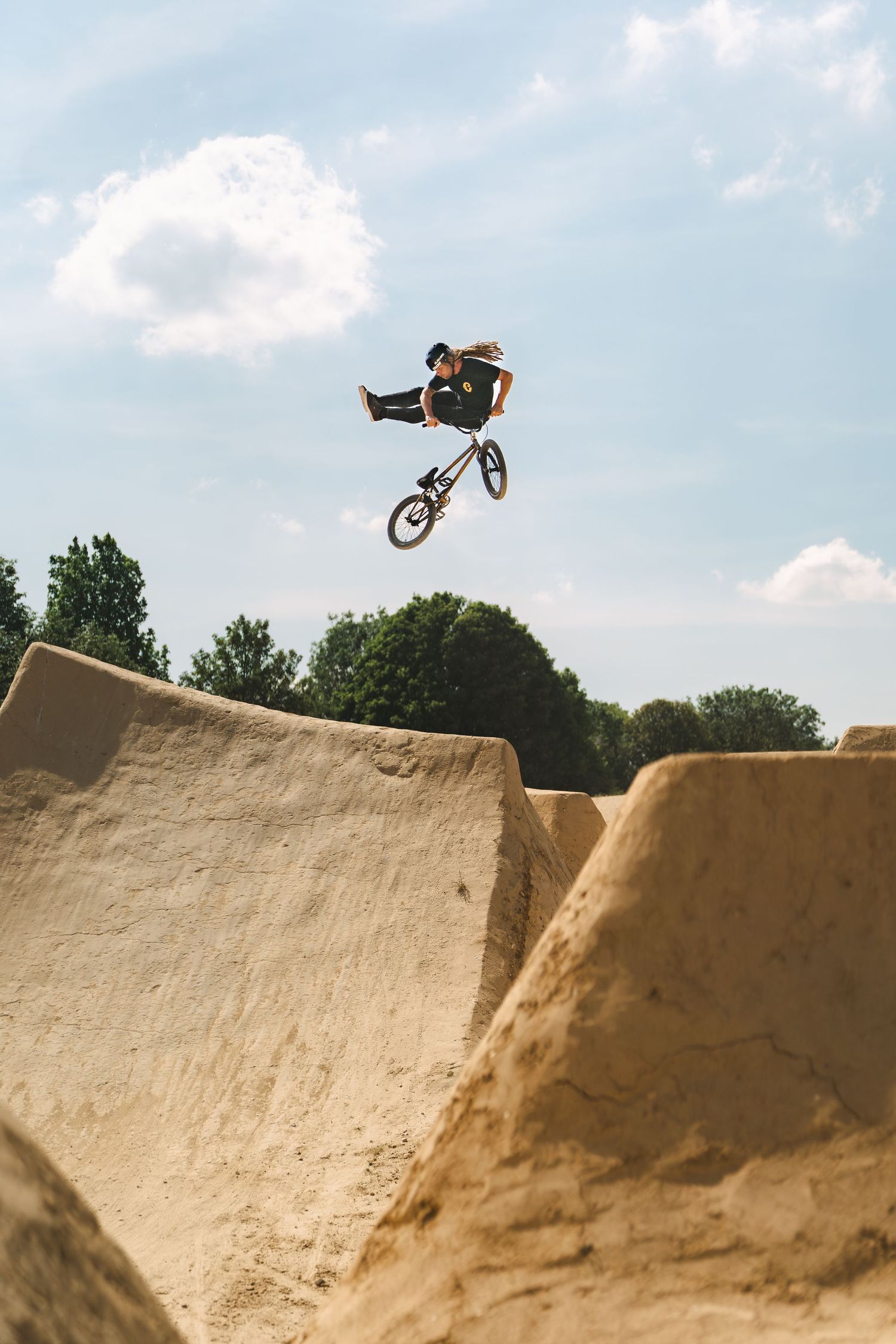 A BMX rider performs a tail whip trick off a large sandy dirt jump under blue skies.