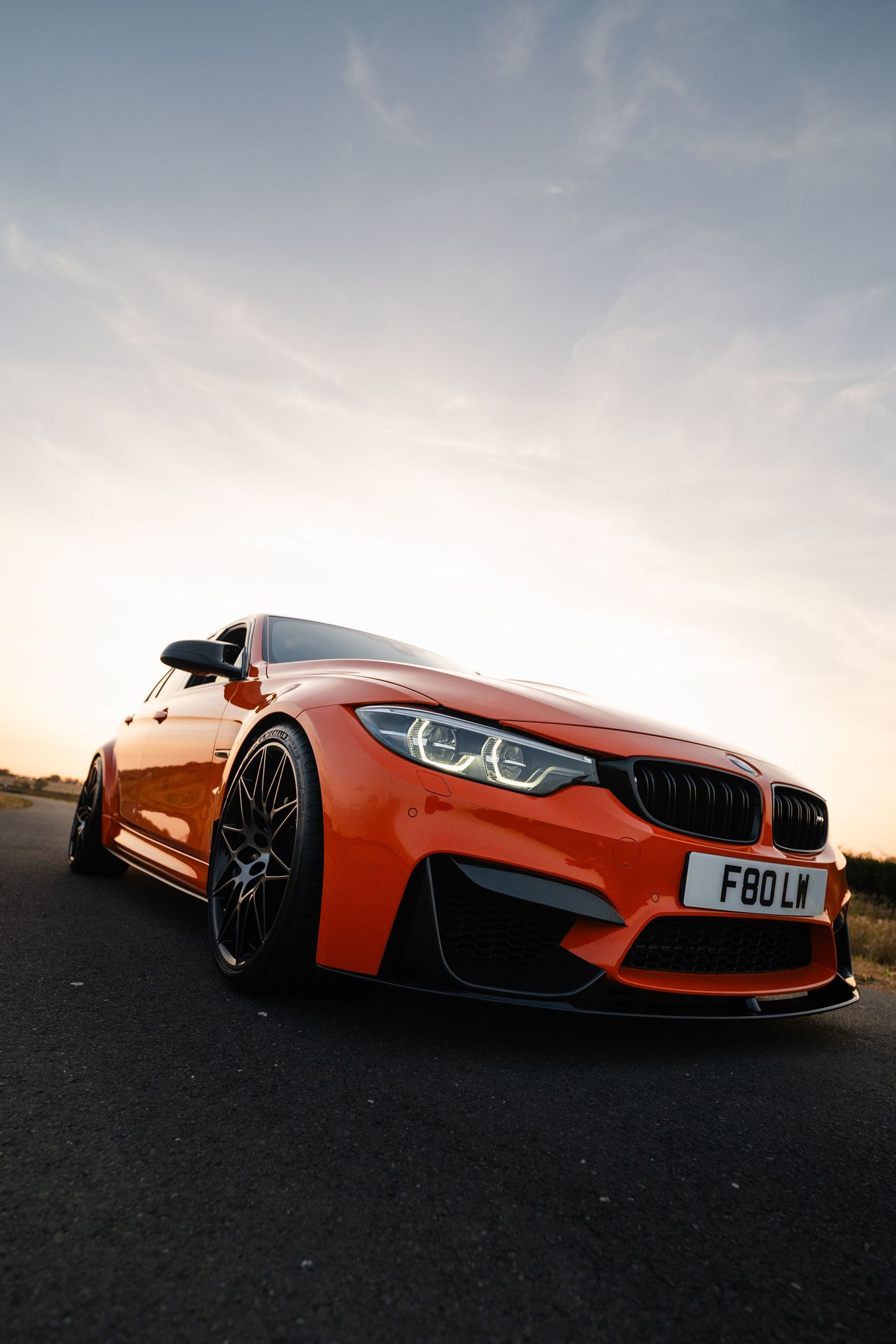 Orange BMW M3 F80 sports car shot from low angle at sunset on empty road with dramatic sky.