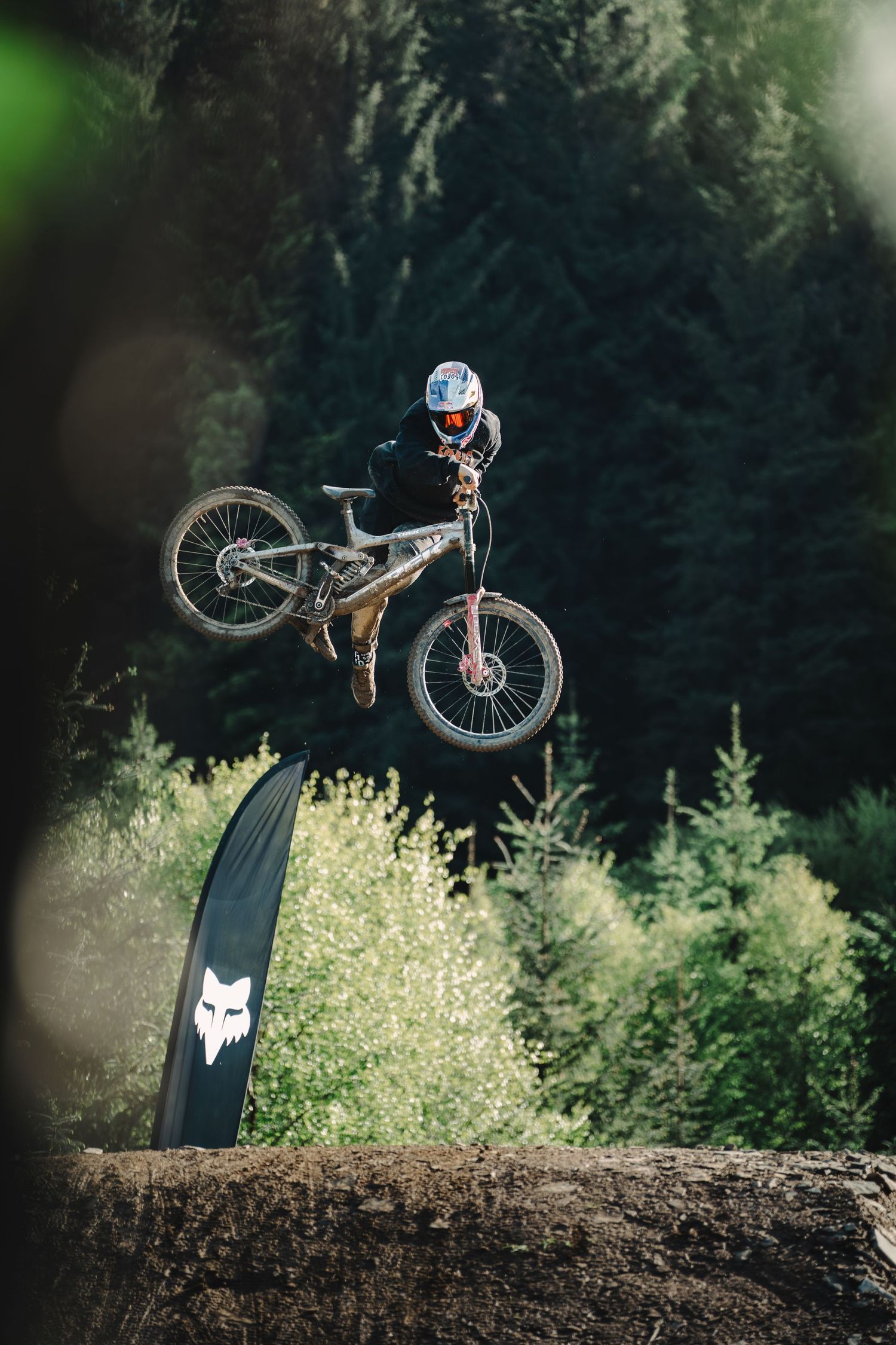 Mountain biker performing a high jump trick over a dirt mound with Fox Racing banner and forest backdrop.