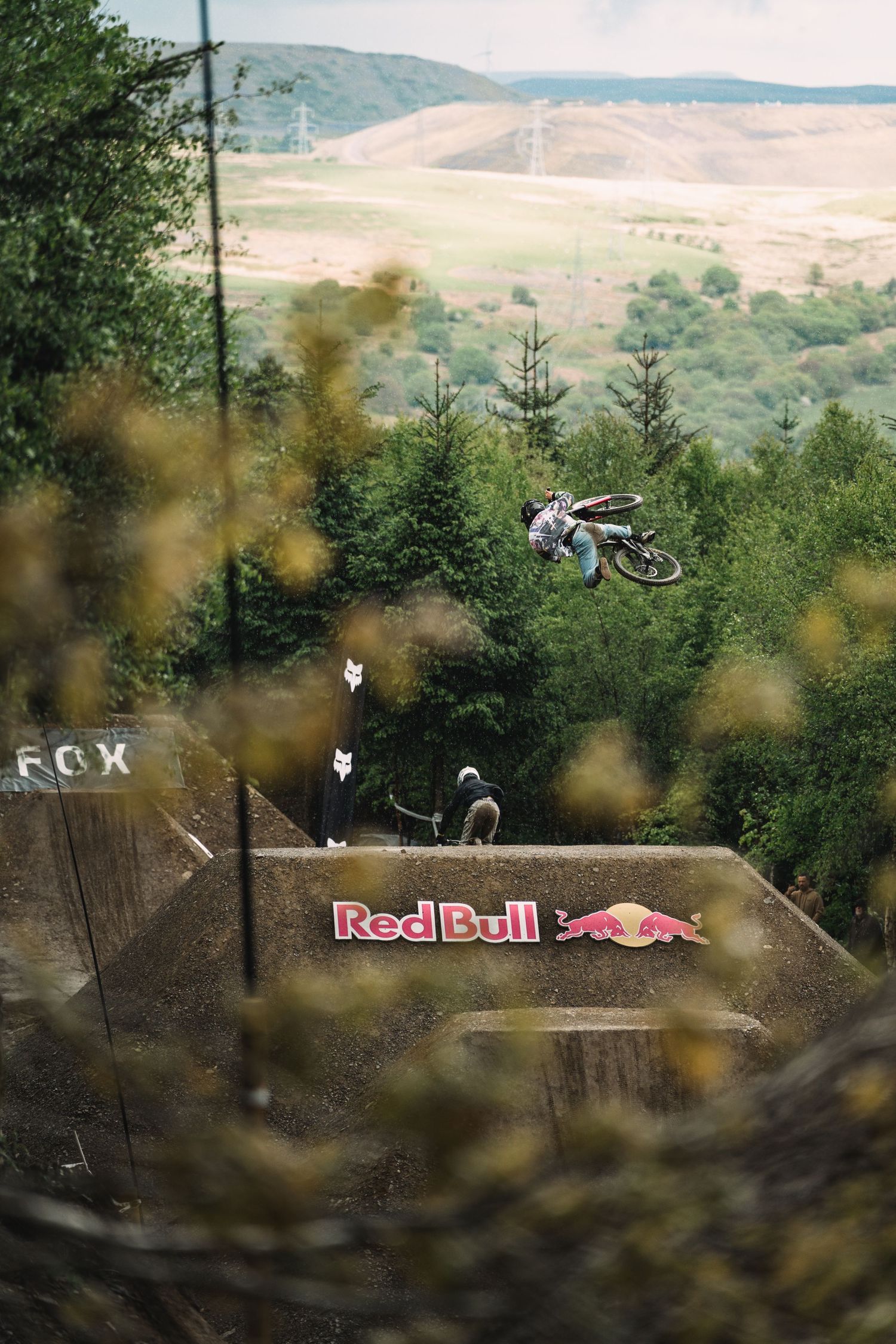 Motocross rider performs aerial trick over a Red Bull branded ramp with forest backdrop.
