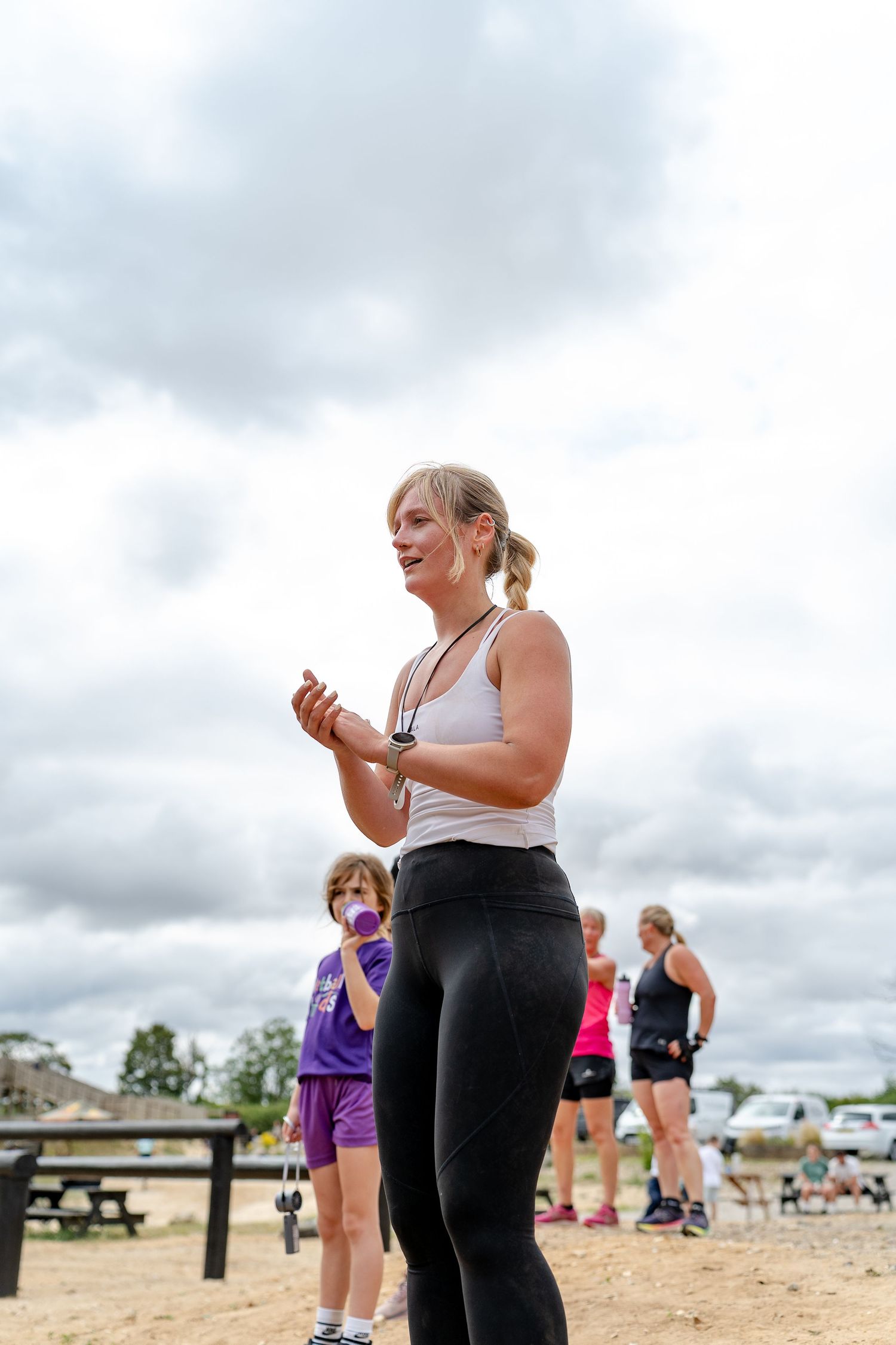 Woman in white tank top and black leggings clapping outdoors at a fitness event under cloudy skies.