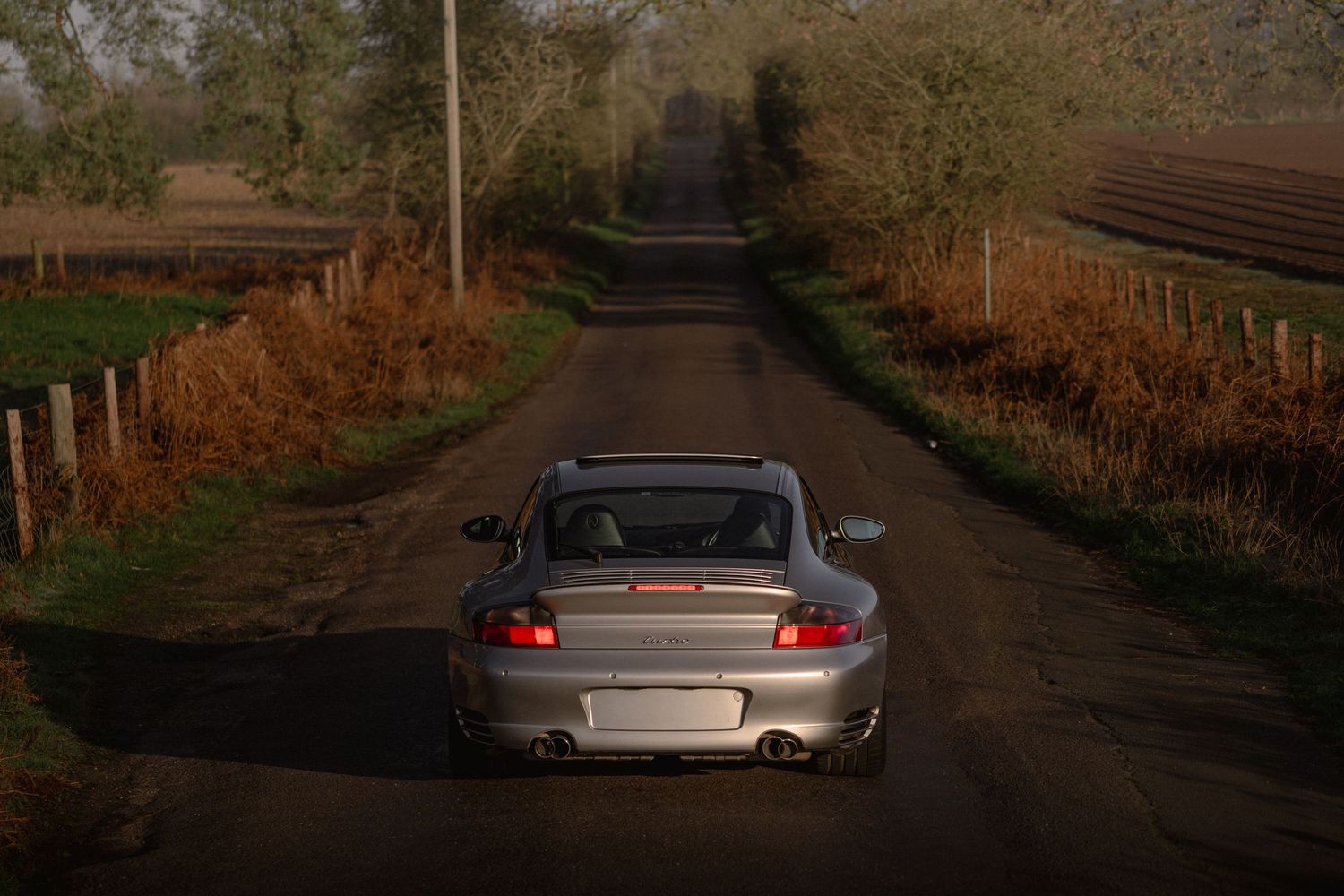 Rear view of a silver Porsche 911 Turbo on a narrow rural road surrounded by autumn hedgerows.