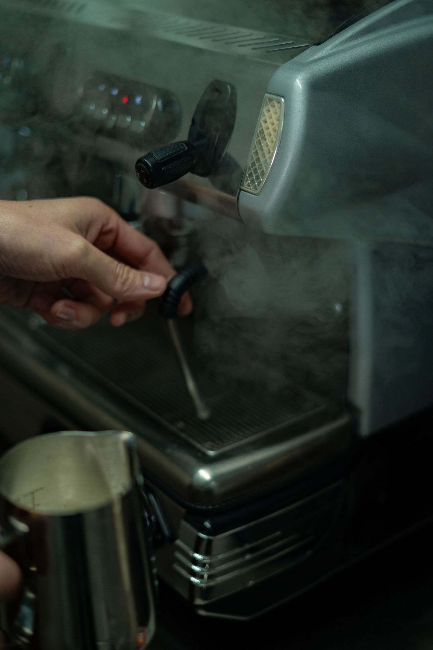 Steam rising from a professional espresso machine steam wand with milk pitcher beside it.